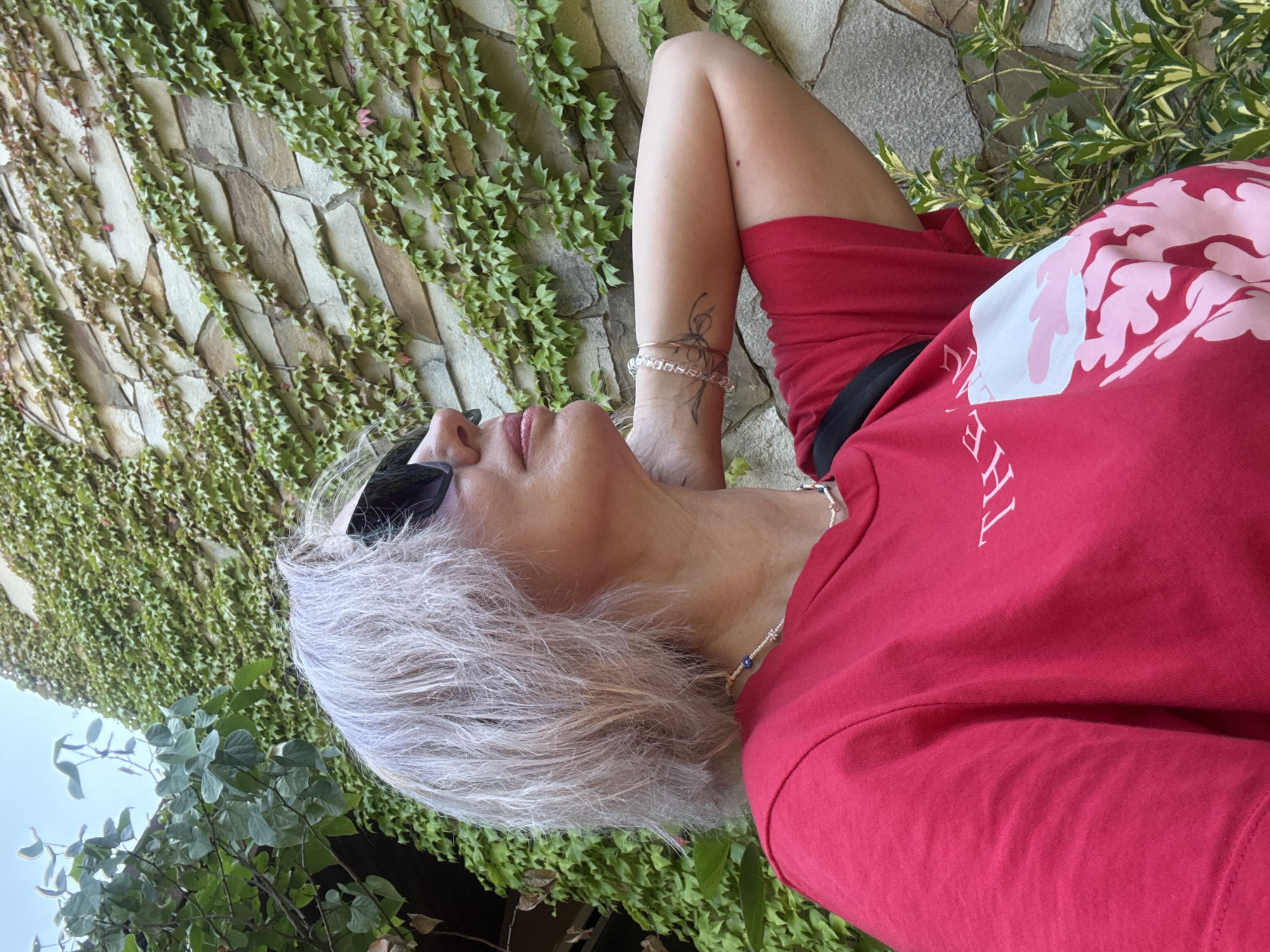 A medium shot captures a woman with short, ash-blonde hair, wearing sunglasses and a red t-shirt with a white and pink graphic design. She is standing outdoors in front of a stone wall covered in green ivy. Her right arm is bent and resting behind her head, and she is looking upwards and to her right. A subtle necklace is visible around her neck, and she has a tattoo on her right arm. The background shows more of the ivy-covered wall and some foliage to the left. The setting appears to be a garden or courtyard, possibly in the city of Estosadok, Russia, given the style of the architecture and vegetation. The lighting suggests it is daytime.