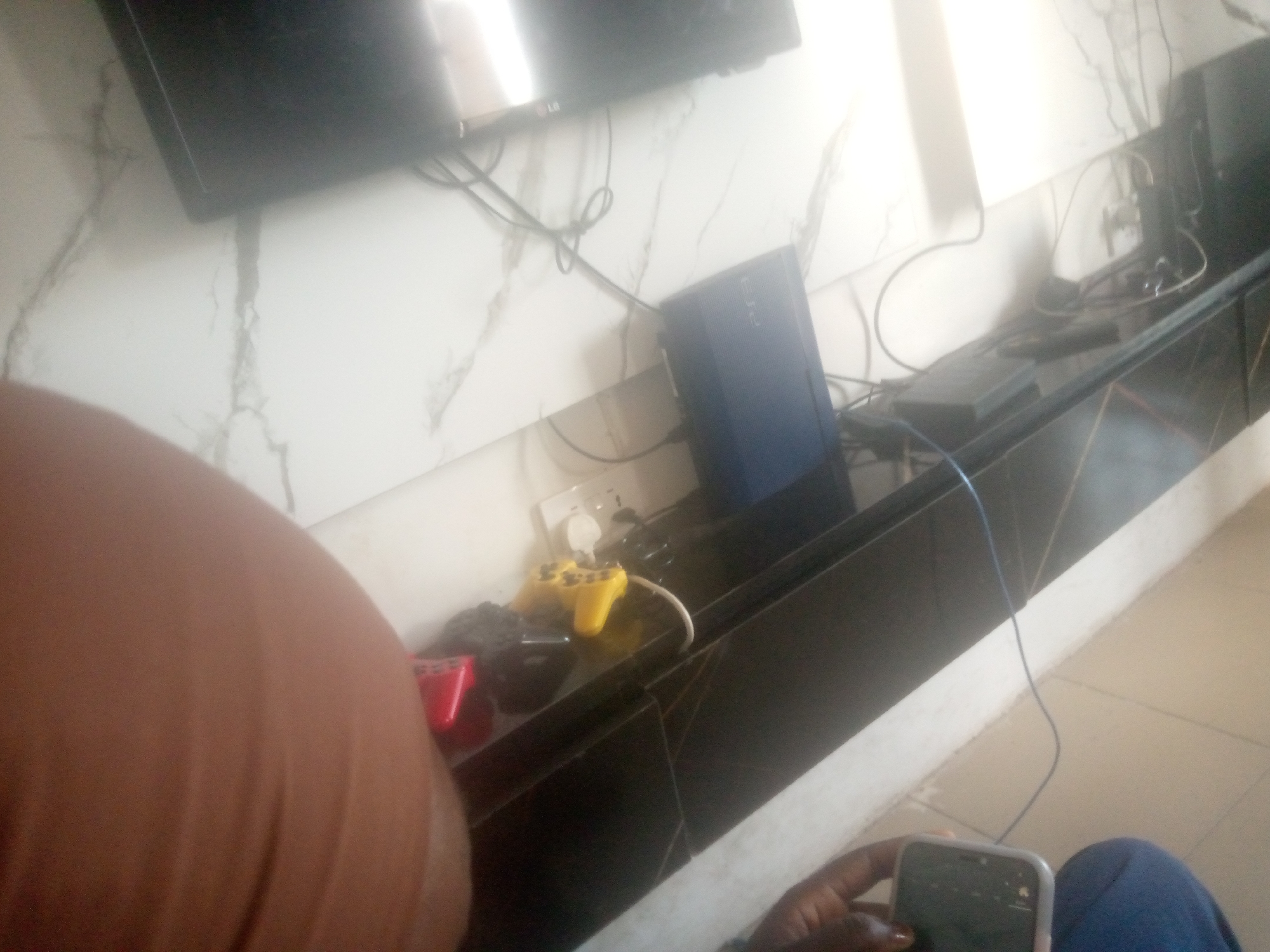 The image depicts an indoor scene, likely a living room or entertainment area, with a prominent television mounted on a wall with a marble-like finish. Below the television, a black entertainment console houses various electronic devices. On the console, a blue PlayStation 3 console is visible, along with a black power strip and several cables. Two video game controllers, one yellow and one red, are also present on the console. A white electrical outlet is mounted on the wall above the console. The foreground of the image is dominated by a blurred brown object, which appears to be a person's body part, possibly an arm or leg, and a hand holding a smartphone. The overall lighting suggests it is daytime, with some glare from an unseen light source. The environment appears to be a typical urban dwelling, but no specific location within Jalingo, Nigeria, can be determined from the visual information alone.