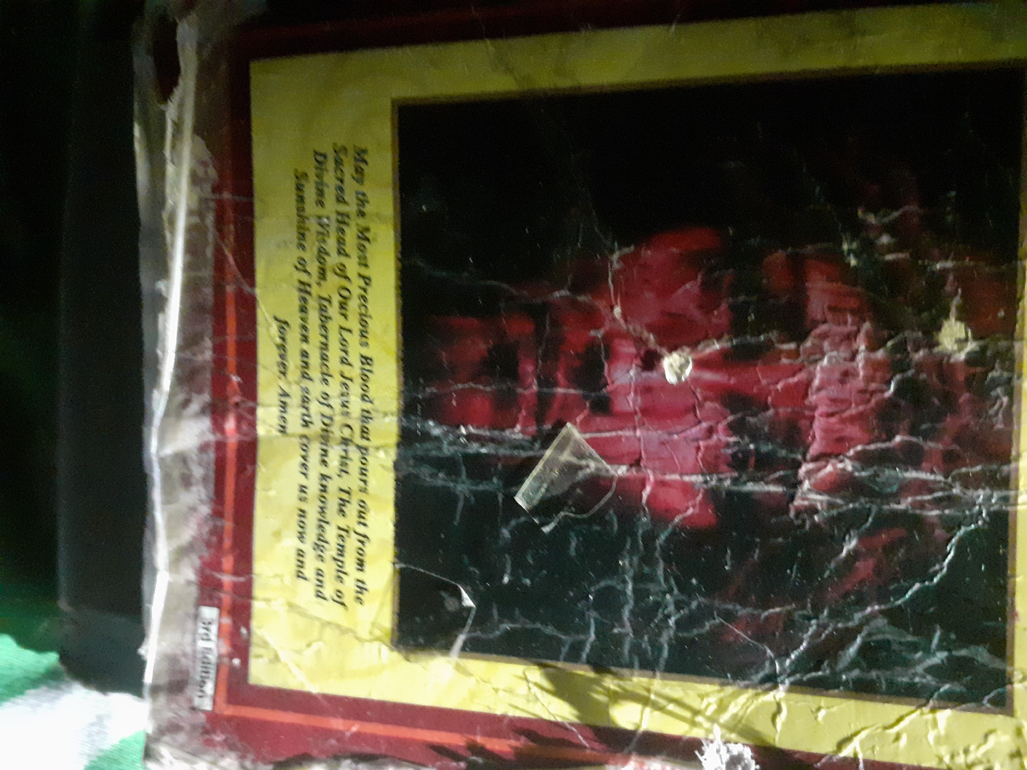 The image shows the back cover of a book with a religious inscription and a dark image with red undertones. The text, written in yellow lettering against a black and red background, reads "May the Most Precious Blood that pours out from the Sacred Head of Our Lord Jesus Christ, The Temple of Divine Wisdom, Tabernacle of Divine knowledge and Sunshine of Heaven and earth cover us now and forever, Amen." At the top left of the cover, "3rd Edition" is visible. The overall condition of the book cover appears worn and damaged, with visible creases and a slightly torn edge. The image does not provide any specific details about the city of Jalingo, Nigeria, or any inhabitants of the city.