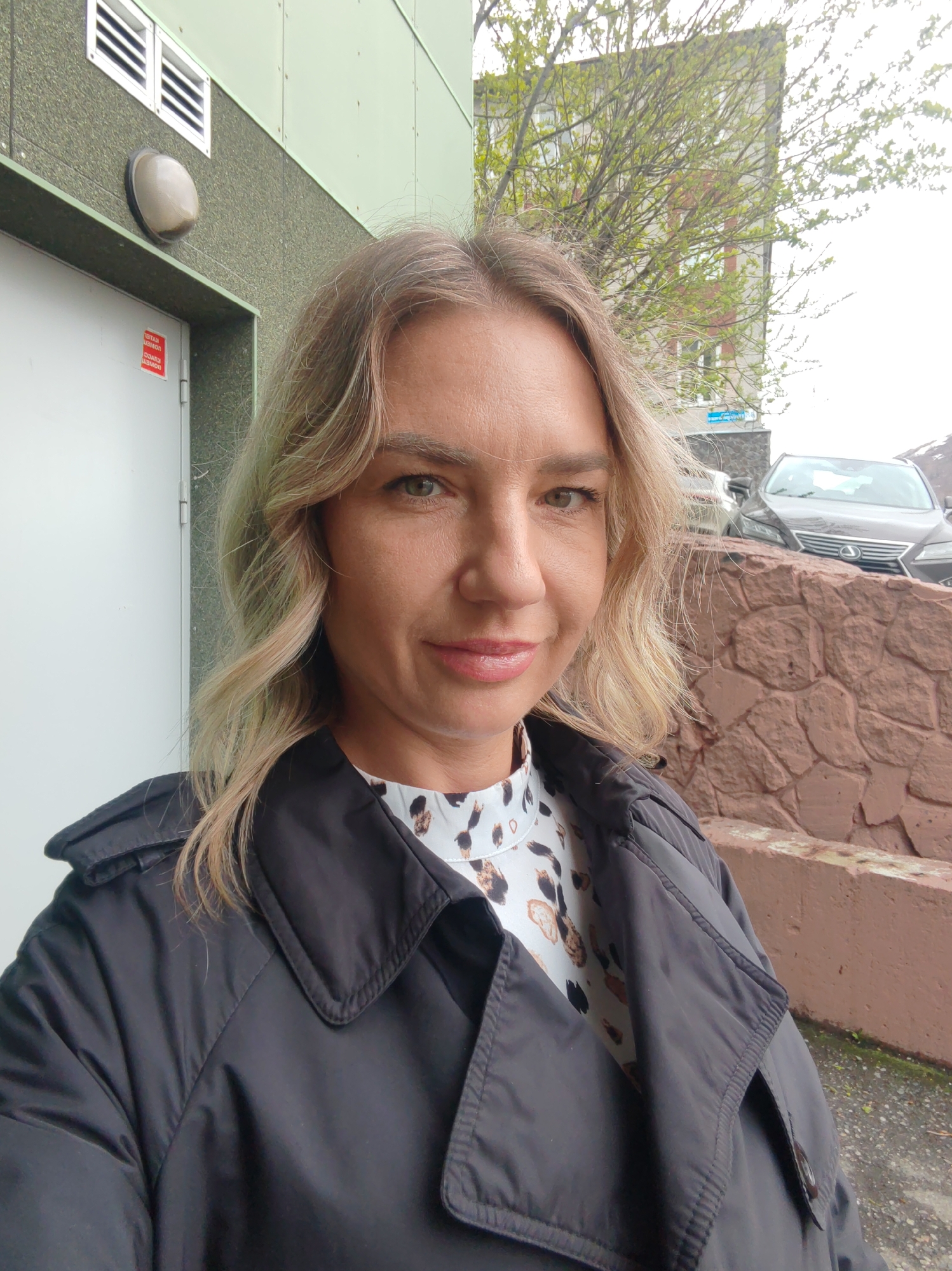 The image shows a woman with blonde hair and a black coat, posing for a selfie. She is standing outdoors in what appears to be a city environment. In the background, there's a building with a green facade and white door. A car is visible behind a stone wall. A street sign with Russian text, indicating "Вулканная" (Vulkannaya Street), suggests the location is Petropavlovsk-Kamchatsky, Russia.