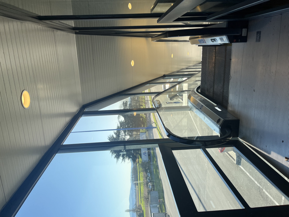 The image displays the interior of a modern building, dominated by an escalator and large windows, captured under bright, sunny conditions. The scene is oriented vertically but would show an escalator descending towards the foreground on the right side of the frame if rotated correctly. The escalator features dark steps and handrails, and a white panel with blue characters, possibly "0000," is visible near its upper section.
To the left, expansive, dark-framed windows offer a clear view of the outside world. Through the glass, a vibrant daytime landscape is visible, including green trees, a road, a parking area, and distant rolling hills or mountains under a bright blue sky. Reflections of the sky and internal elements, including what appears to be a person's silhouette, are present on the windowpanes.
The building's interior features a light-colored, paneled ceiling with multiple circular, recessed light fixtures emitting a warm light. The walls are similarly paneled. The flooring around the escalator is a dark grey. No people are actively using the escalator, and the scene is static, suggesting a quiet moment. The overall impression is of a spacious and well-lit environment, blending modern architecture with an open view of nature. The context places the location in Dorno, Italy.