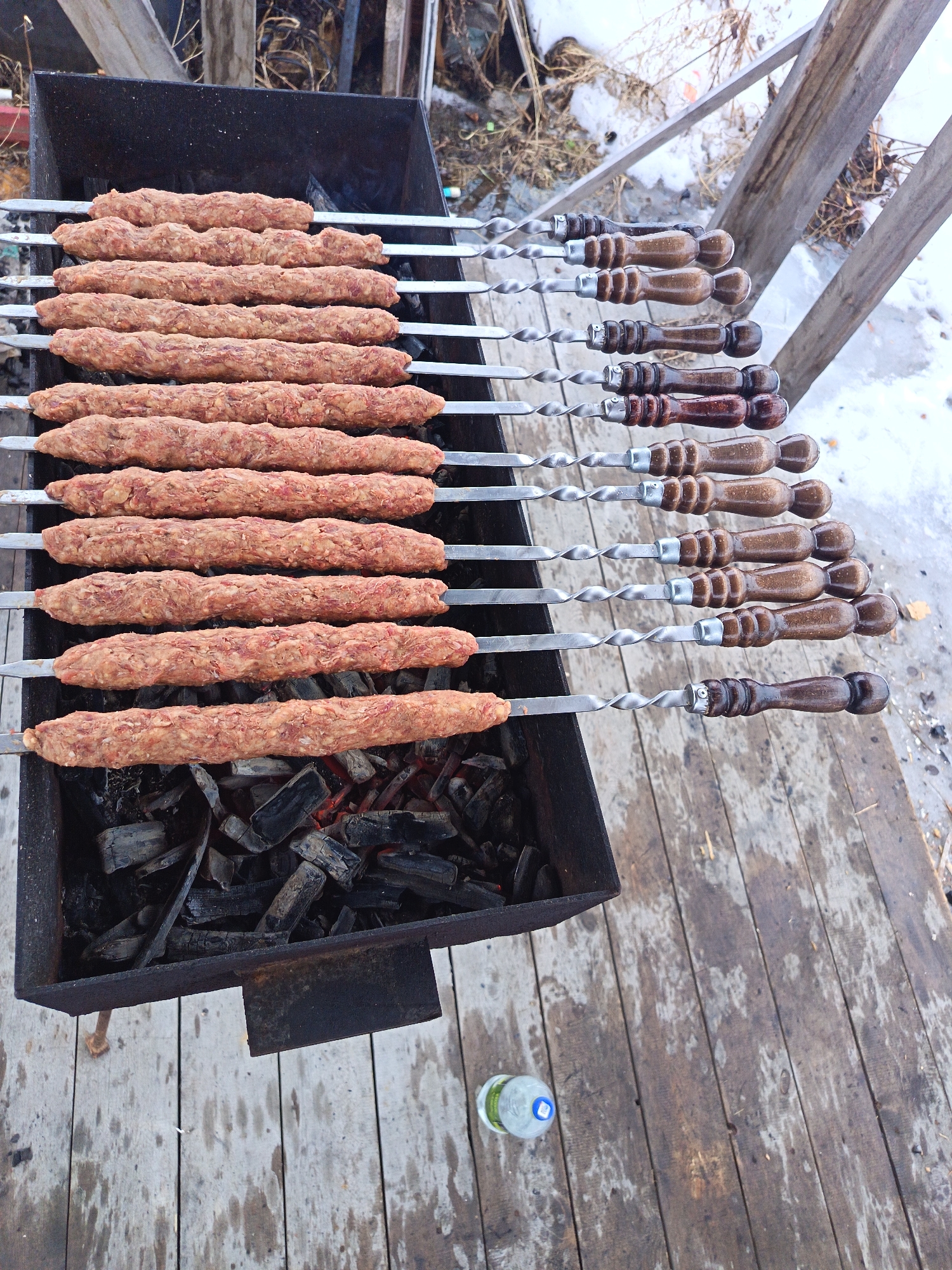 A top-down medium shot captures a barbeque scene. A rectangular metal barbeque grill is filled with hot coals. Eleven skewers, each bearing a long, cylindrical portion of raw meat, are arranged across the grill. The skewers have metal shafts and wooden handles.
The barbeque grill is situated on a weathered wooden deck. Patches of snow are visible between the wooden planks of the deck. A partially visible water bottle sits at the lower right of the frame, on the deck.
Based on visual markers like building styles, the image may have been taken in Nizhnevartovsk, Russia. No people are visible in the image. The action visible is raw meat placed over hot coals on metal skewers, implying a barbeque cooking activity.