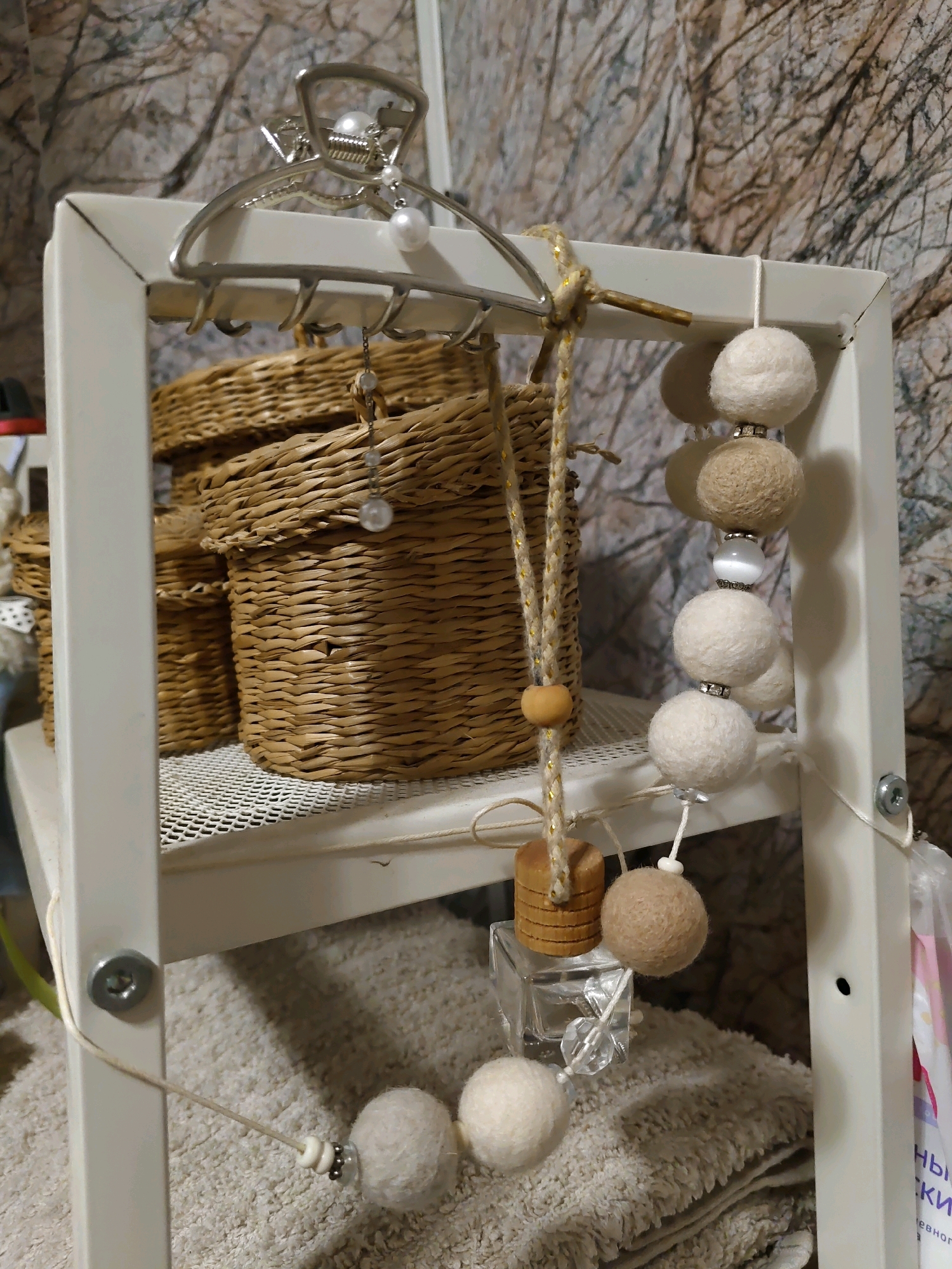 In Rostov-on-Don, Russia, a white metal shelf unit holds several items. On the top shelf, a large wicker basket is partially visible, and a smaller wicker basket is placed in front of it. A silver claw clip with pearl embellishments hangs from the top bar of the shelf unit. Below the top shelf, another row of wicker baskets is visible. A string with felt balls and beads hangs from the top bar, extending down to the second shelf and then down to the bottom shelf. On the second shelf, next to the string of felt balls, is a small square glass object with a wooden cylindrical piece attached to it. The bottom shelf contains a folded textured towel, and another string with felt balls and beads hangs from the shelf unit, resting on the towel. The background consists of a textured wall.