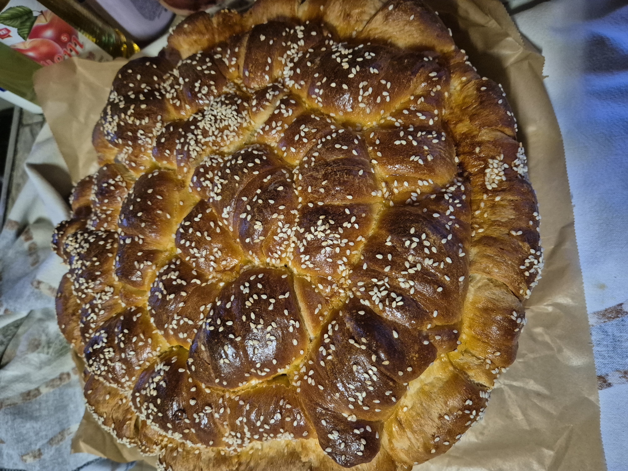 A close-up, overhead shot showcases a golden-brown, round loaf of challah bread generously sprinkled with sesame seeds. The bread has a braided or twisted appearance with several distinct rolls or sections clustered together. The texture is glossy, suggesting a rich egg wash. It is placed on a piece of brown parchment paper, which in turn rests on a patterned fabric. In the background, out of focus, are a few bottles, one of which has an apple label. The lighting is warm and highlights the browning of the bread. The scene appears to be indoors, likely in a kitchen or dining area.