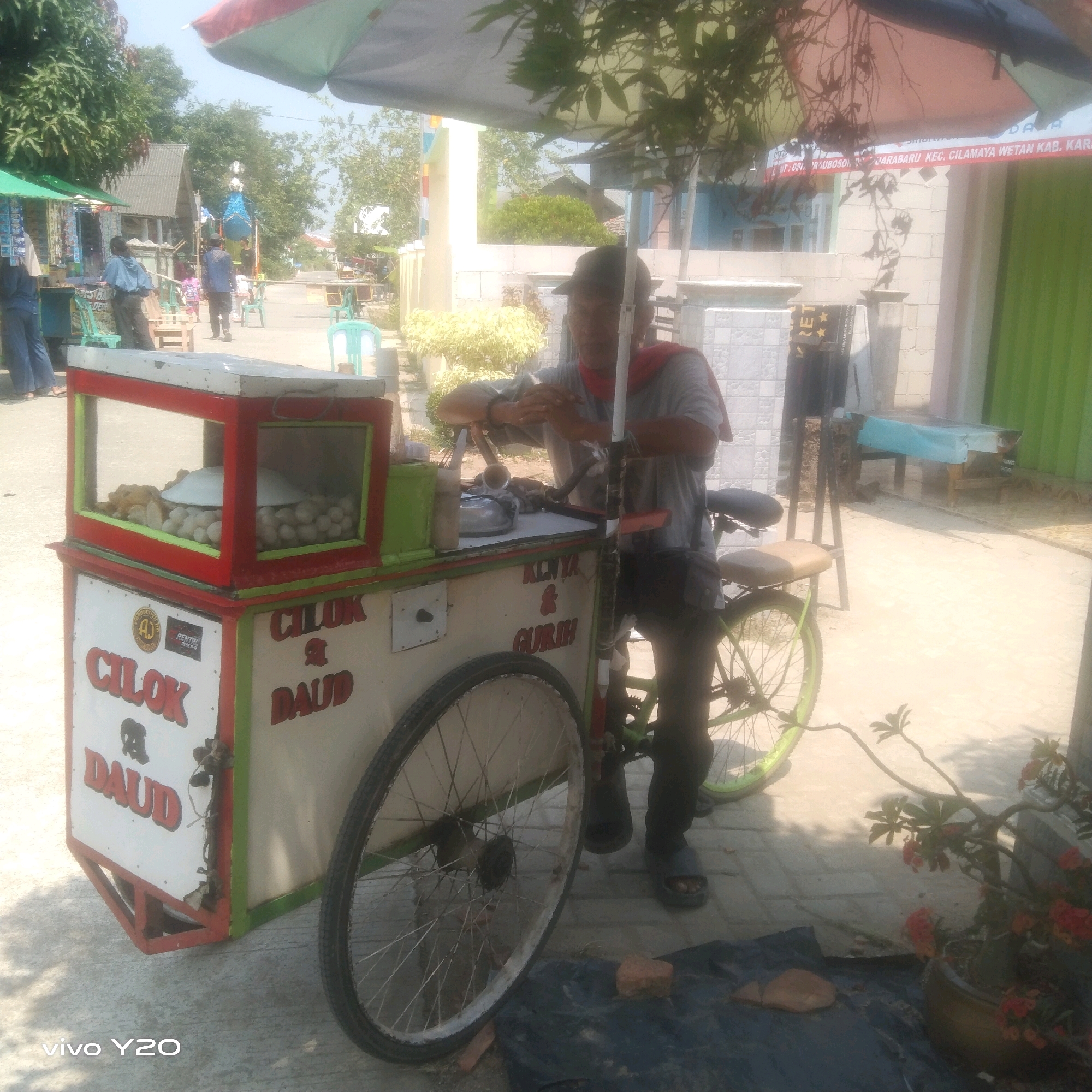 An outdoor, eye-level, medium shot captures a street vendor preparing food from a mobile cart. The cart, painted red, white, and green, displays the words "CILOK & DAUD" and "KENYAL & GURIH" in red lettering. Inside a glass-enclosed compartment, white spheres, likely the food being sold, are visible. A man, wearing a gray shirt and a red cloth around his neck, stands behind the cart, operating it. He has a dark-colored hat and is actively engaged in his task. The cart is attached to a bicycle, indicating its mobile nature. The background reveals a street scene with other vendors, buildings, and greenery, suggesting a typical urban marketplace environment. The lighting indicates it is daytime, with shadows cast by the umbrella above and surrounding objects.