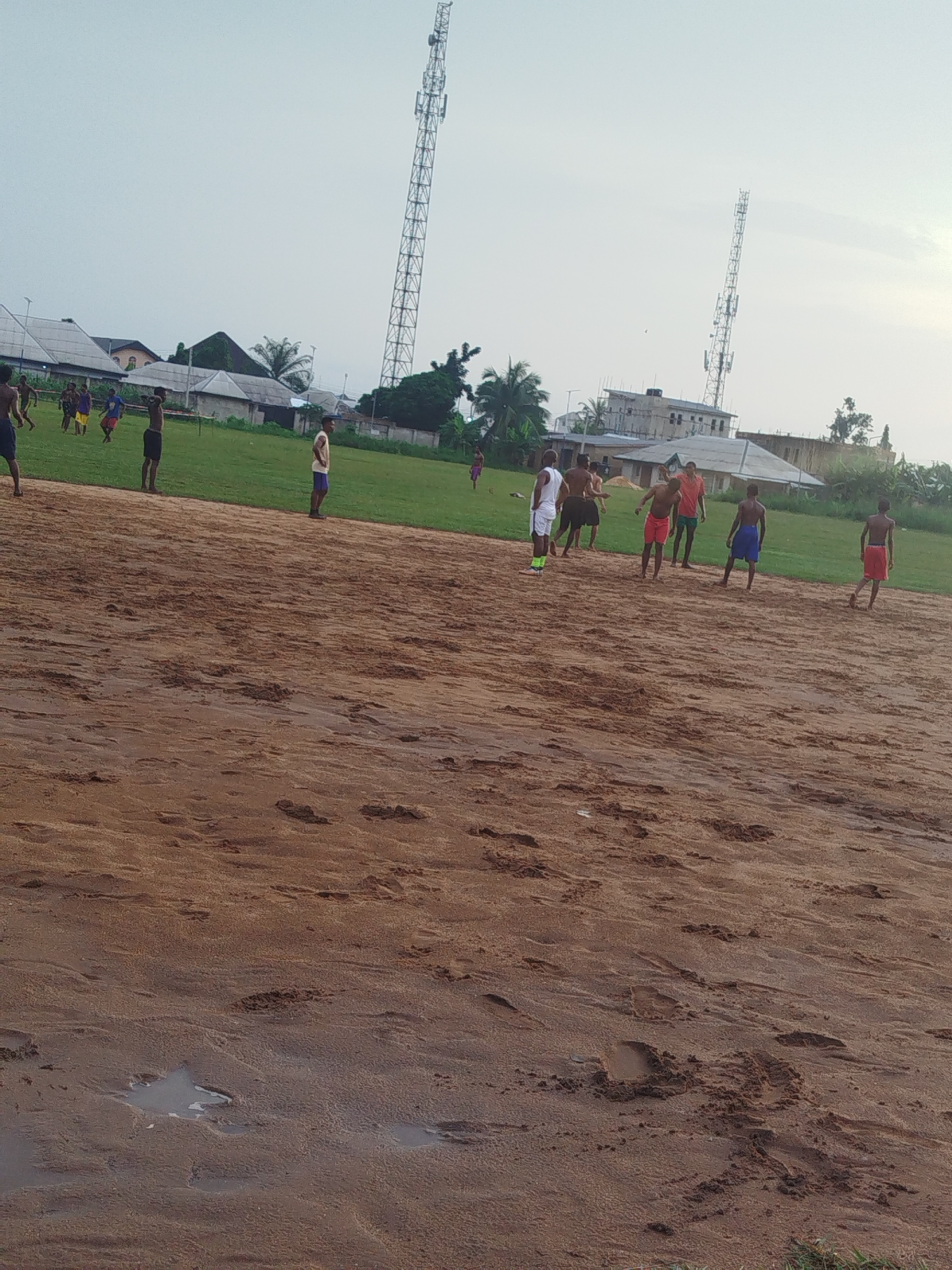 The image depicts a field, possibly in Ikot Mbonde, Nigeria, indicated by the presence of tall telecommunication towers in the background, a common infrastructure element in urban settings. In the foreground, the ground is predominantly brown, sandy, and uneven, with evidence of footprints and puddles of standing water. A group of young men are scattered across the field. Most are shirtless, with some wearing shorts of various colors including red and blue. One man wears a white shirt and white shorts with green socks. They appear to be engaged in an informal sporting activity. In the background is a green grassy area with houses and vegetation, as well as a portion of a building under construction with exposed brick. The sky is overcast.