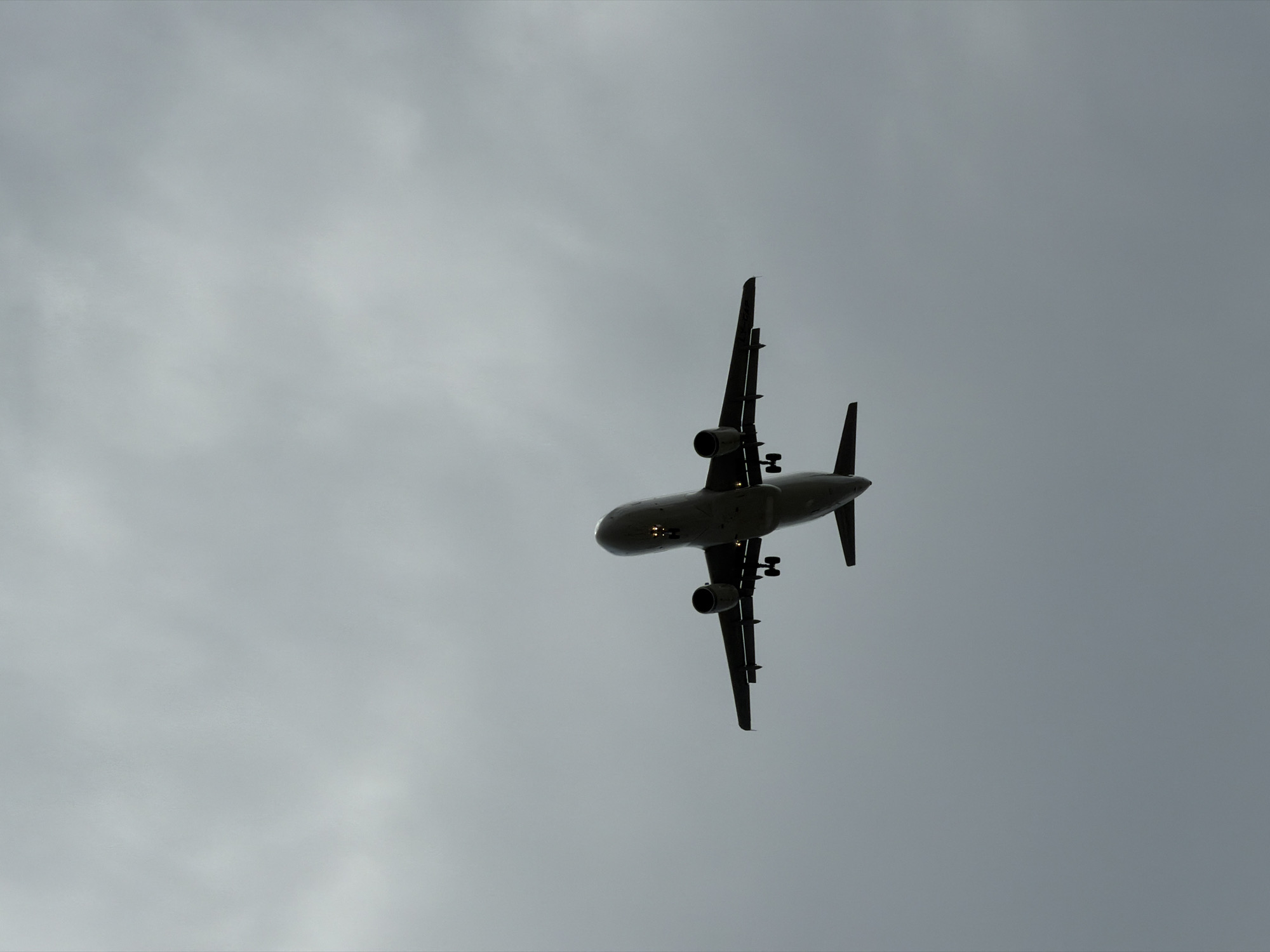 An airplane is captured in silhouette against a heavily overcast sky, seen from below. The aircraft is descending, with its landing gear deployed and illuminated by its own lights. The underside of the plane, including its wings, fuselage, and tail, is clearly visible. The image has a monochromatic feel due to the gray sky and the dark silhouette of the plane. The specific location in Burgas, Bulgaria, cannot be determined from the provided image alone; it simply shows an airplane in the sky.