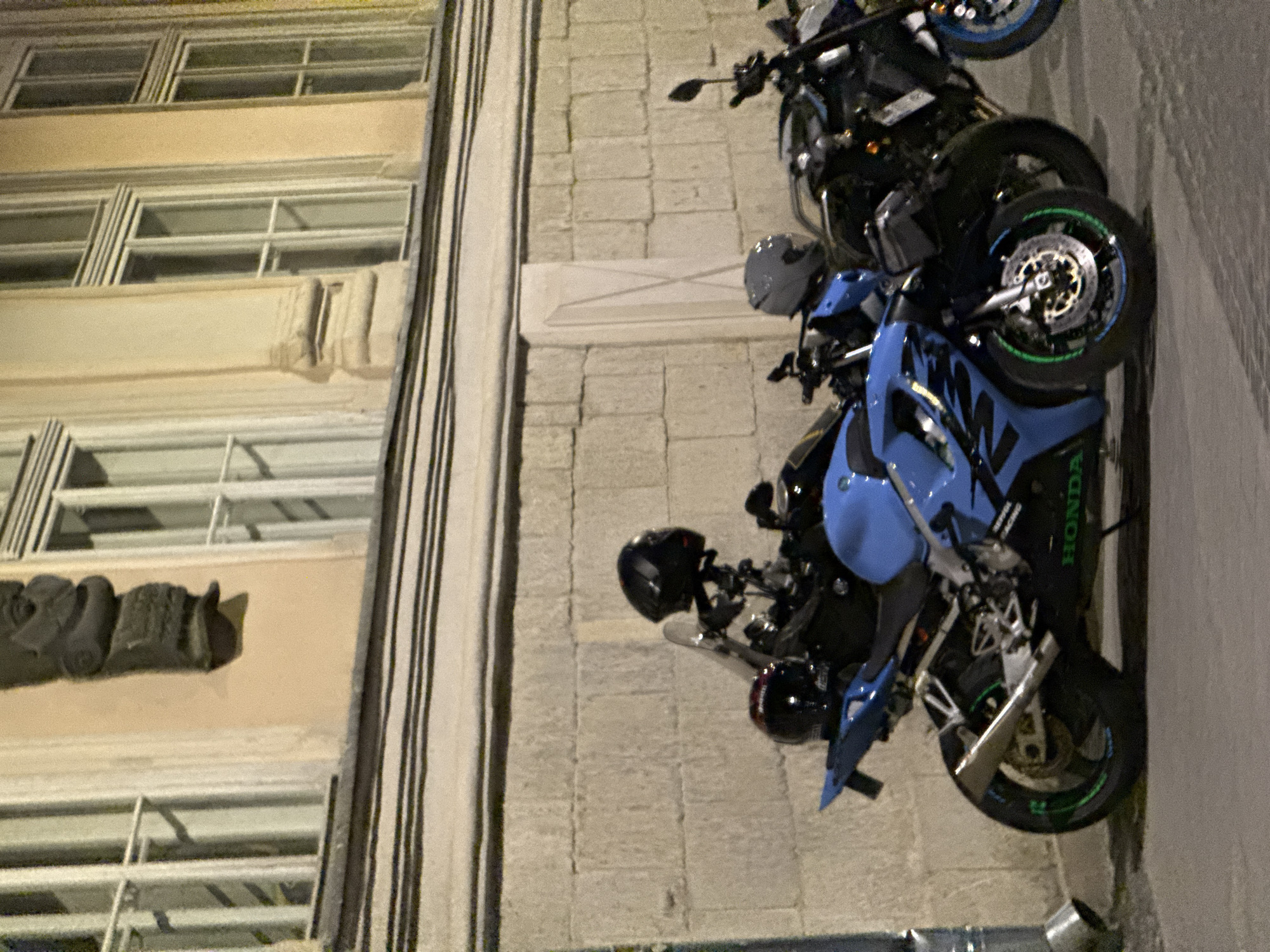 This is a medium-distance, eye-level, vertical, low-light photograph of several parked motorcycles in front of a building. The building features a stone facade with large, multi-paned windows and ornate architectural details, including a decorative carving above the windows. The motorcycles are positioned on a paved surface, likely a sidewalk or street.
The most prominent motorcycle in the foreground is a bright blue sportbike with black accents. It has a silver exhaust pipe and green detailing on the wheels. The word "HONDA" is visible in green lettering on the lower fairing. A black helmet rests on the seat of this motorcycle, and another black helmet is attached to the handlebars.
Behind the blue motorcycle, other motorcycles are visible. One appears to be a dark-colored sportbike with a black helmet on its seat. Further back, another dark motorcycle is partially obscured. The scene suggests a common urban setting where motorcycles are parked.
The overall impression is that of an undisturbed, static scene. There are no people clearly visible in the image, and no apparent activity is taking place. The lighting indicates it is either dusk or nighttime. Based on the architectural style and the overall ambiance, this scene could be located in the historic city center of Lviv, Ukraine, a city known for its well-preserved architecture.