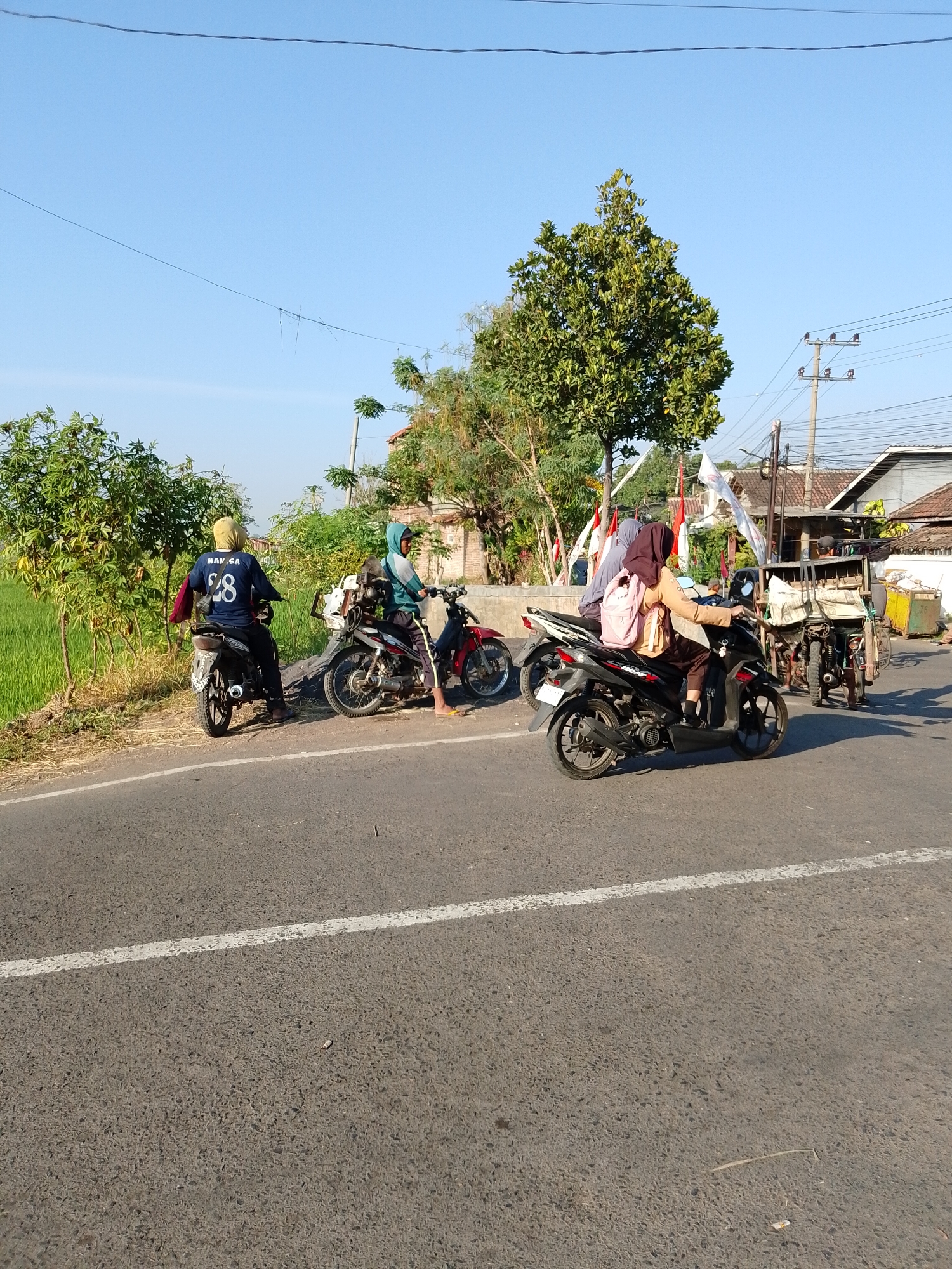 The image depicts a street scene in Pasuruan, Indonesia, on a clear, sunny day. The road is asphalt with a white painted line. On the side of the road, there are several motorcycles and people.
In the foreground, a woman wearing a brown hijab and a light brown jacket is riding a black motorcycle. She has a pink backpack on. To her left, a man in a teal hijab and jacket is on a red and black motorcycle. Behind him, a person in a blue shirt with the number "28" on the back is seated on a black motorcycle, facing away from the camera.
On the right side of the image, a man in a green hijab and jacket is standing next to a motorcycle with a cart attached. The cart appears to be carrying various items covered in white cloth. Further back, there are buildings with terracotta roofs and telephone poles with wires.
The environment includes lush green vegetation, including rice paddies and trees, flanking the road. The sky is a clear, bright blue. The overall impression is of a quiet, rural or semi-rural road with daily life activities.