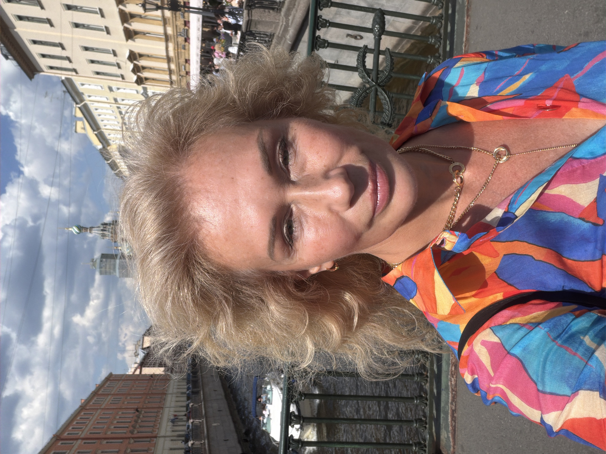 A blonde woman with curly hair smiles softly, looking directly at the camera in a close-up, outdoor selfie. She wears a brightly colored top featuring a bold pattern in shades of blue, orange, pink, and yellow, along with a delicate gold necklace.
The background reveals an urban setting, identified as Saint Petersburg, Russia, characterized by classical European architecture lining a canal or river. A decorative dark green metal railing of a bridge is prominent next to her. The sky is partly cloudy, suggesting bright daylight, likely midday or early afternoon. In the distance, other buildings, including one with a faint onion-domed spire, are visible. The water of the canal or river can be seen below, with what appears to be a boat and distant figures. The woman seems relaxed and content. No visible text is present.