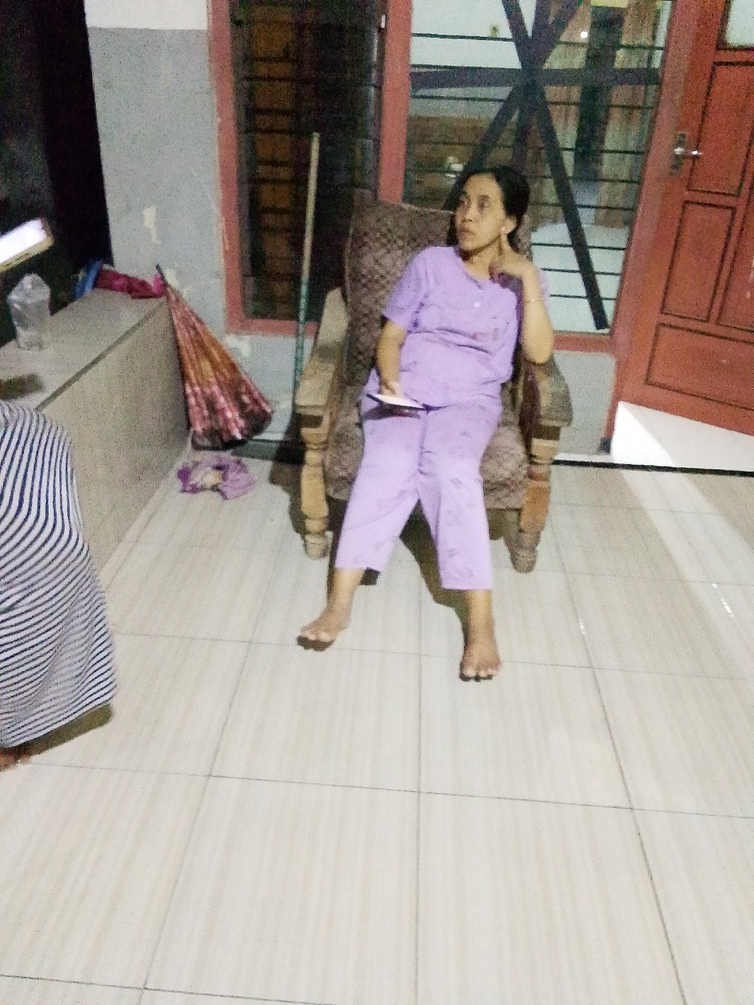 A woman in a light purple pajama set is seated in a wooden chair indoors, holding a small white object in her left hand. She is looking to her right with her right hand resting on her chin. The room appears to be a residential interior, possibly in Bulurejo, Indonesia, with tiled flooring and a wall with a pink-trimmed window on the right. To the left of the woman, a portion of another person's striped clothing is visible. Various objects are scattered in the background, including a folded umbrella, a plastic cup, and what appears to be a dark piece of fabric. A wooden pole stands behind the woman.