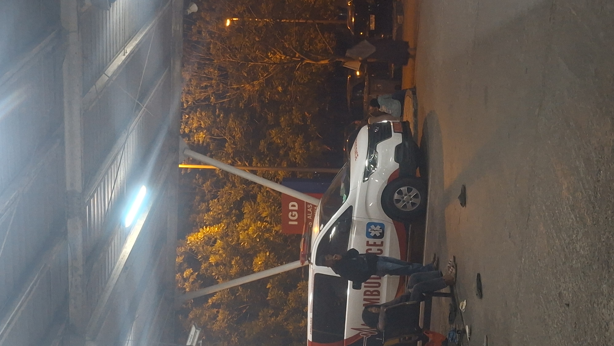 An ambulance with "AMBULANCE" text on its side is parked under a covered outdoor area. A man in a blue jacket and jeans stands by the ambulance, and a person with dark hair sits on a bench to the left of the ambulance. Several pairs of sandals are scattered on the ground. In the background, trees are visible, and a sign with "IGD" in red letters and other text is mounted on a pole. The scene appears to be outside a medical facility in Banda Aceh, Indonesia.