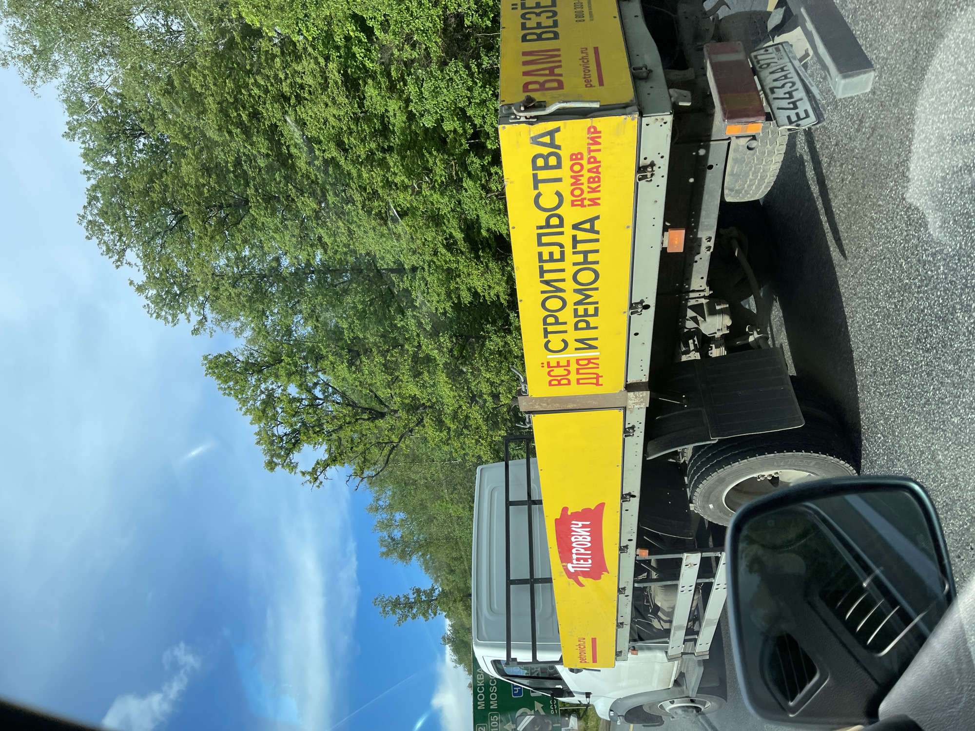 The image shows a view from inside a vehicle, focusing on a truck alongside on the road. The truck has a yellow advertising banner for "Petrovich," a construction and renovation supply company. The banner reads "Всё для строительства и ремонта домов и квартир" (Everything for construction and renovation of houses and apartments) and "Вам Везёт" (You're Lucky). The company's website, petrovich.ru, and phone number, 8 800 333-, are also displayed. The truck's license plate number is E443AA 977.
In the background, there are trees and a blue sky with some clouds. A road sign indicates "Москва 2 (Moscow 2)" and "105 MOSCO," suggesting the location is near Moscow.
Based on the license plate's regional code "977", the vehicle is registered in Moscow. The road sign indicating "Moscow 2" indicates a proximity to the city. The proximity to Moscow in the image aligns with the description of the city "Serp I Molot" as the city is also in Russia.