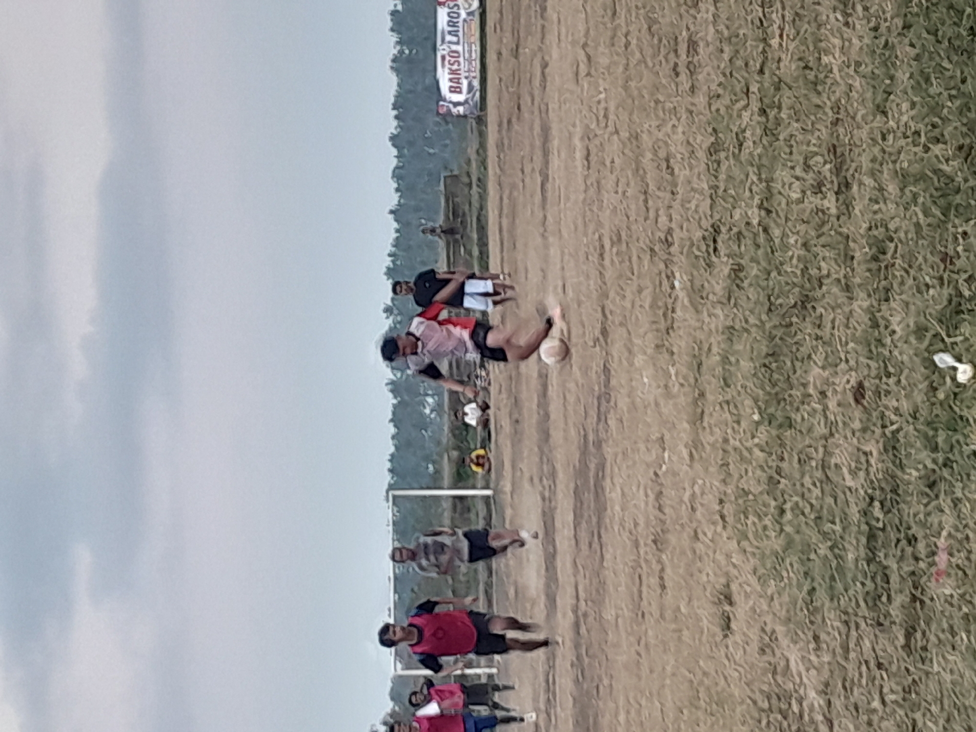 This image depicts a football game on a dirt field in Genteng, Indonesia. The sky is overcast with a pale blue hue. A group of young men are actively participating in the game. One individual in the foreground is mid-action, kicking a soccer ball. He is wearing a white and red jersey with black shorts. Several other players are visible on the field, some running and others standing. A football goalpost with a white frame is situated in the background. To the right of the field, a banner with text, partially obscured by the low resolution, reads "BAKSO LAROS." The terrain is uneven and dry, with sparse patches of grass. The surrounding environment appears to be rural, with trees and foliage visible in the distance.
