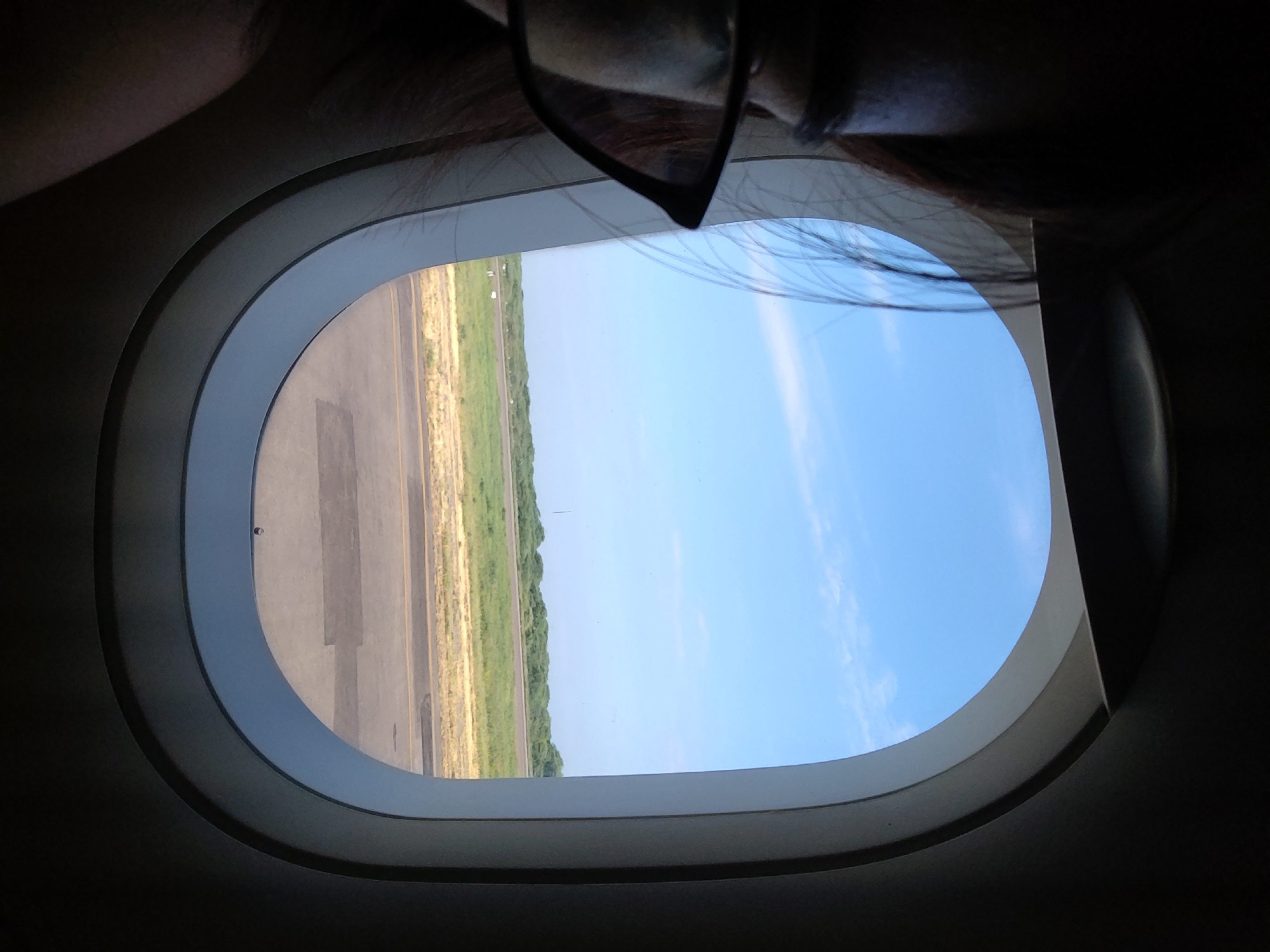 A view from an airplane window shows a portion of an airport runway with grass and trees alongside a portion of clear blue sky with wispy clouds. A pair of eyeglasses and a strand of hair are partially visible in the foreground, suggesting the viewer is inside the plane looking out the window. The airplane window is oval-shaped with a white frame and a dark interior around it. The image was likely taken in the city of Tumbes, Peru.