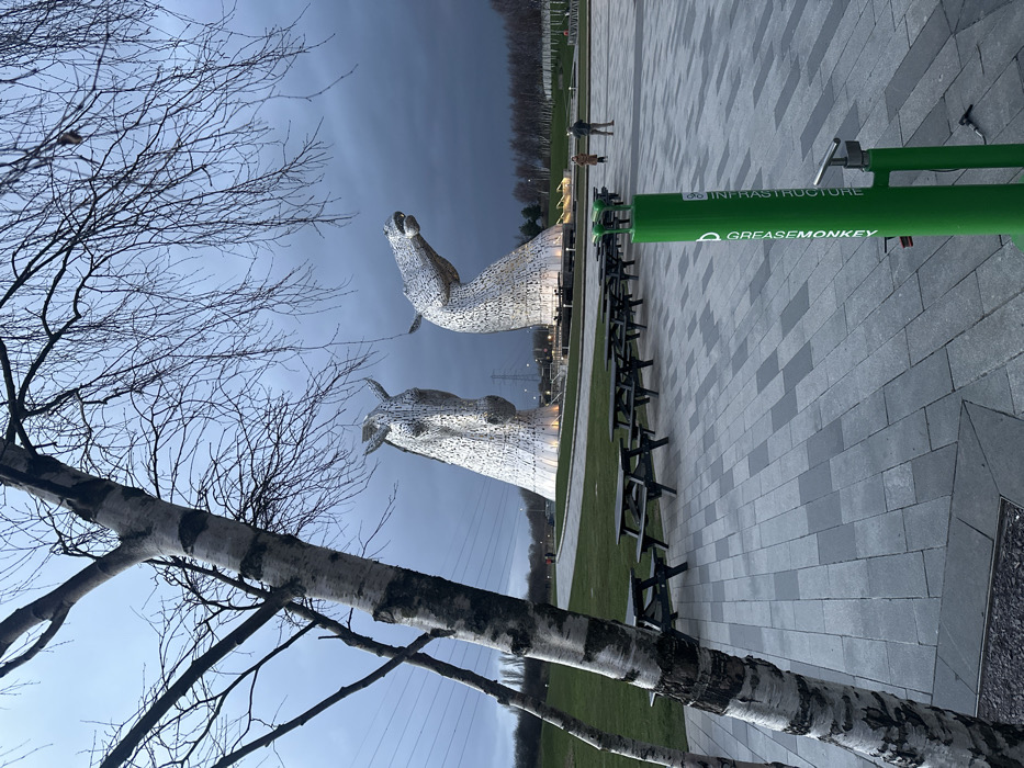 The image depicts an outdoor scene at dusk or early evening, centered on two large, illuminated equine head sculptures known as The Kelpies, located in Helix Park, Falkirk, United Kingdom. The sky is heavily overcast with dark grey clouds.
In the foreground, on the left, a silver birch tree with stark, bare branches extends diagonally from the lower left towards the top right, partially obscuring the left sculpture. Its trunk exhibits characteristic white and dark grey patterned bark. On the right side of the foreground, a bright green, cylindrical bicycle repair station stands upright, featuring "GREASEMONKEY" and "INFRASTRUCTURE" text printed vertically, along with a pump attached.
The midground consists of a paved area made of rectangular light and dark grey blocks, stretching from the bottom of the frame towards the background. On this paved surface, a linear arrangement of at least seven dark wooden picnic tables with attached benches is visible, extending towards the sculptures. To the left of the paved area, a neatly mown green lawn covers the ground.
The two prominent Kelpies sculptures, constructed from interwoven metallic plates, are situated in the background. The left sculpture presents a horse's head looking mostly forward, while the right sculpture depicts a horse's head in a rearing pose, looking upwards and to the right. Both sculptures are illuminated from their bases by warm artificial lights. Several indistinct human figures are visible walking on a path behind the picnic tables and near the bases of the sculptures. In the far background, power lines and a large pylon are discernible against the horizon, with a line of bare deciduous trees below.