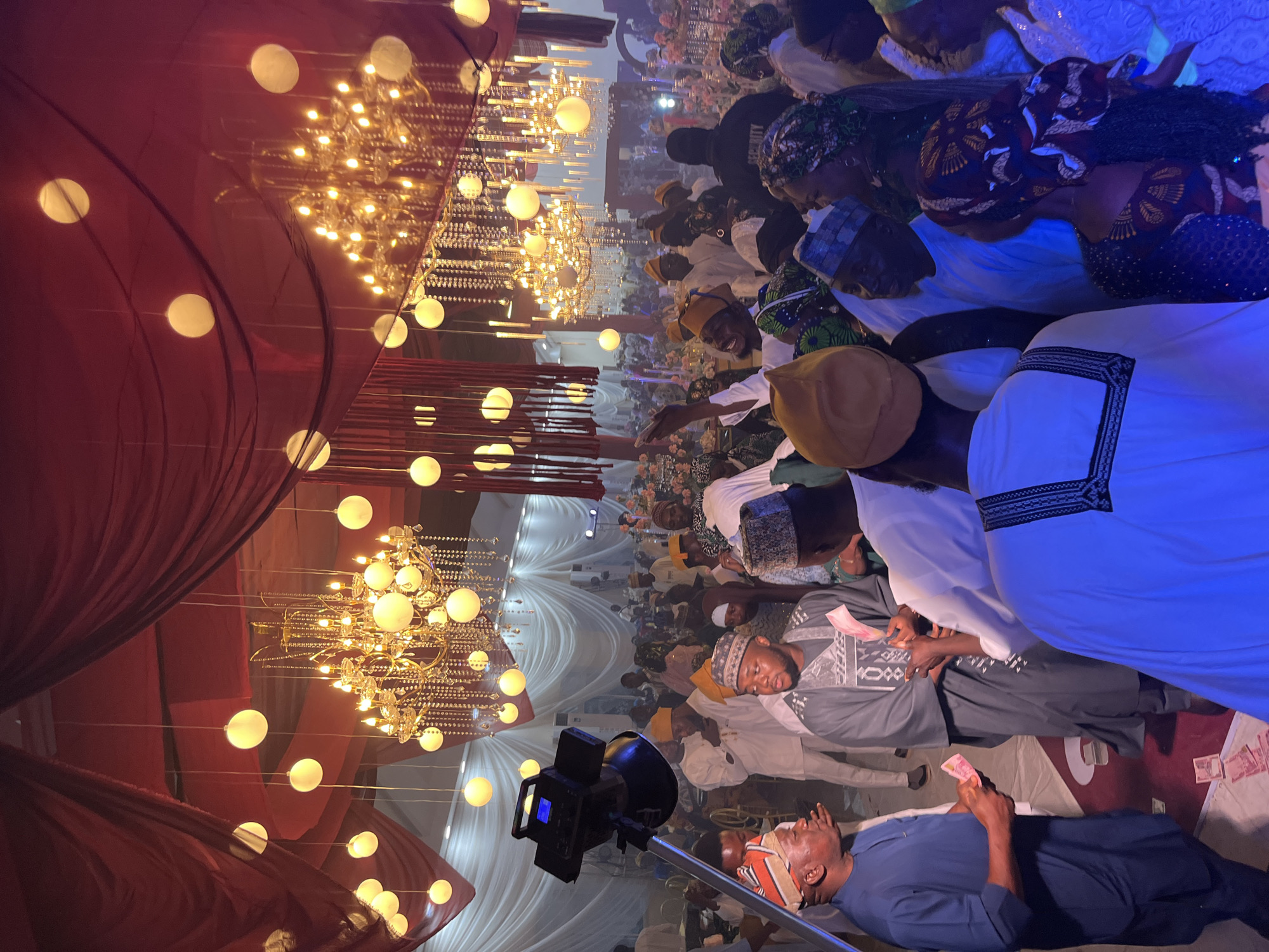 An indoor event appears to be taking place, possibly a wedding reception, with elaborate decorations including red draped fabric and several ornate chandeliers. The room is lit by these chandeliers and numerous round, glowing orb lights. In the foreground, several men are gathered, some in traditional attire, and at least one man in a blue garment is holding cash. Another man in a grey outfit is also holding cash. Further into the background, a large crowd of people, also in formal and traditional clothing, are visible, suggesting a significant gathering. A security guard is also present in the background, identified by a "SECURITY" vest. The overall atmosphere suggests a festive and well-attended occasion.