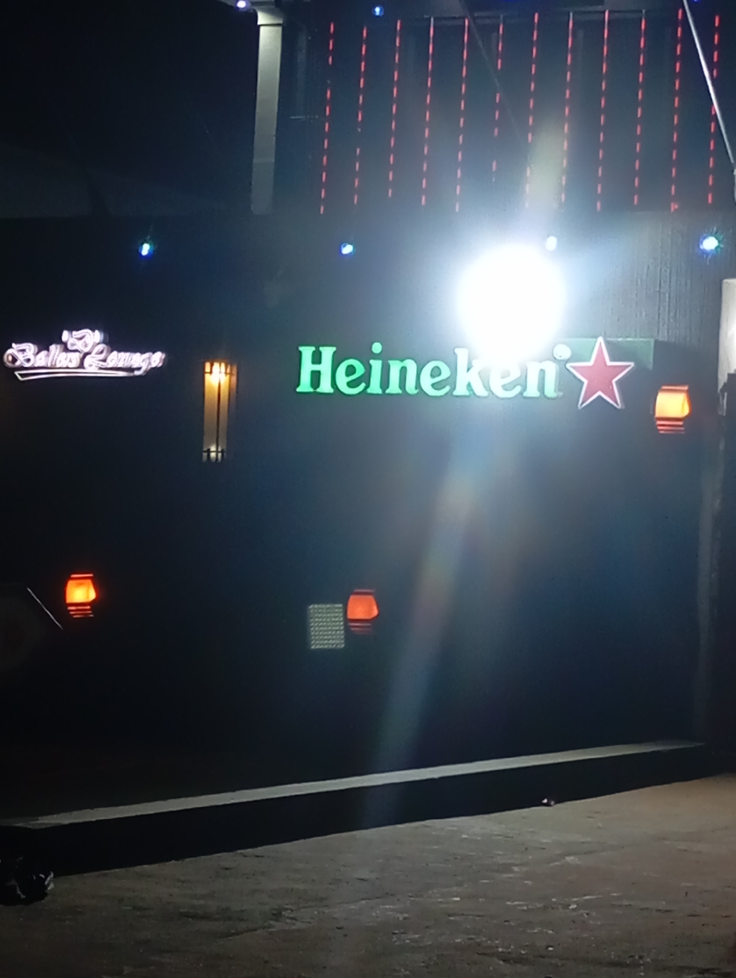 The image depicts a nighttime exterior scene of a commercial establishment in the city of Unknown, Unknown. The scene is dominated by various illuminated signs and lighting fixtures against a dark building facade.
In the center-right, a prominent backlit sign displays the word "Heineken" in green lettering, accompanied by a red star logo to its right. Directly above this sign, an intense light source creates significant lens flare and overexposure, obscuring details in that area. Above the Heineken sign, on the building's upper facade, vertical lines of red dashed lights are visible, likely decorative string lights. Several small, bright blue lights are also scattered across the upper section of the building.
On the left side of the image, positioned higher than the ground level, a white neon-style sign spells "Ballus Lounge" in a script font. Between the "Ballus Lounge" sign and the "Heineken" sign, a tall, narrow, vertically striped wall sconce emits an orange-yellow light. Two similar, but shorter and wider, rectangular wall sconces are visible on the lower walls: one on the far left and another on the far right, both emitting an orange-yellow glow.
The building's facade appears dark and possibly textured. A dark, flat, raised concrete ledge or barrier runs horizontally across the lower middle portion of the image. The ground in the foreground is dark, uneven, and appears to be pavement or concrete. In the bottom-left corner, on the ground near the raised ledge, a small, dark object resembling a pair of shoes or a small bag is present. No individuals are visible in the scene.