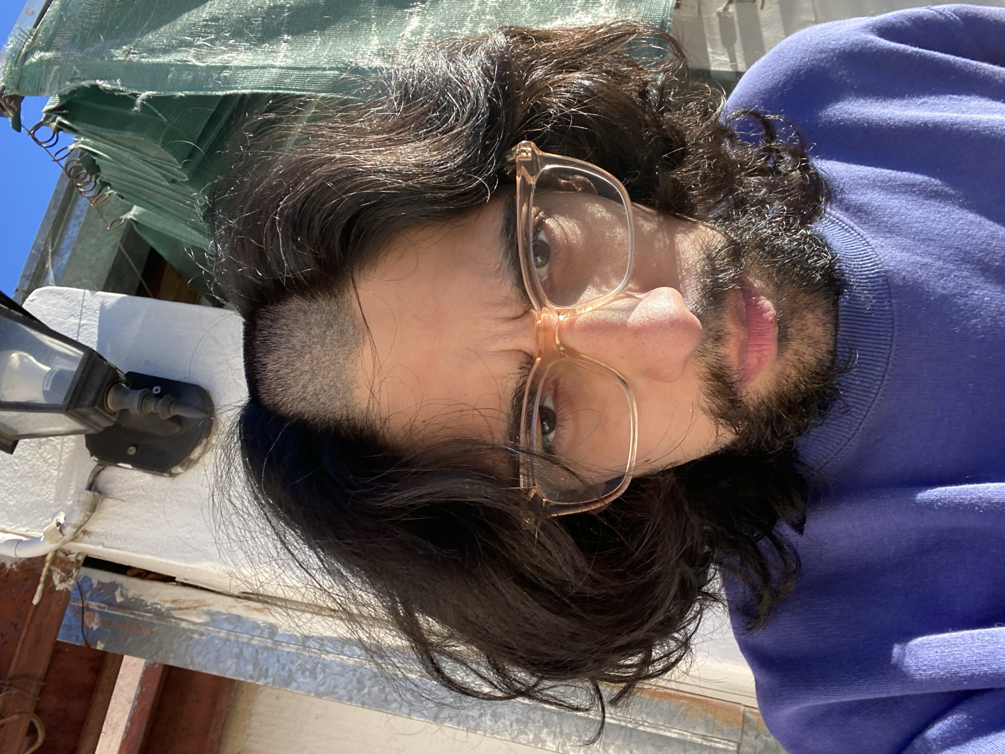 The image shows a person with long, dark, wavy hair, a beard, and light-framed glasses, looking directly at the camera. They are wearing a purple crew-neck sweatshirt. A shaved undercut is visible on the side of their head, and a silver hoop earring is present in their left ear.
The scene is set outdoors during the daytime under a clear, sunny sky, evidenced by the bright natural light and strong shadows cast on the person's face and clothing. In the background, there's a segment of a bright blue sky. A piece of green fabric, possibly an awning or sunshade, held by metal springs, is visible in the upper left. Below this, a black outdoor lamp is mounted on a white textured wall. Further structural elements like wooden beams and metal panels are discernible, suggesting an outdoor area such as a patio, balcony, or rooftop in Buenos Aires, Argentina. No visible text is present.