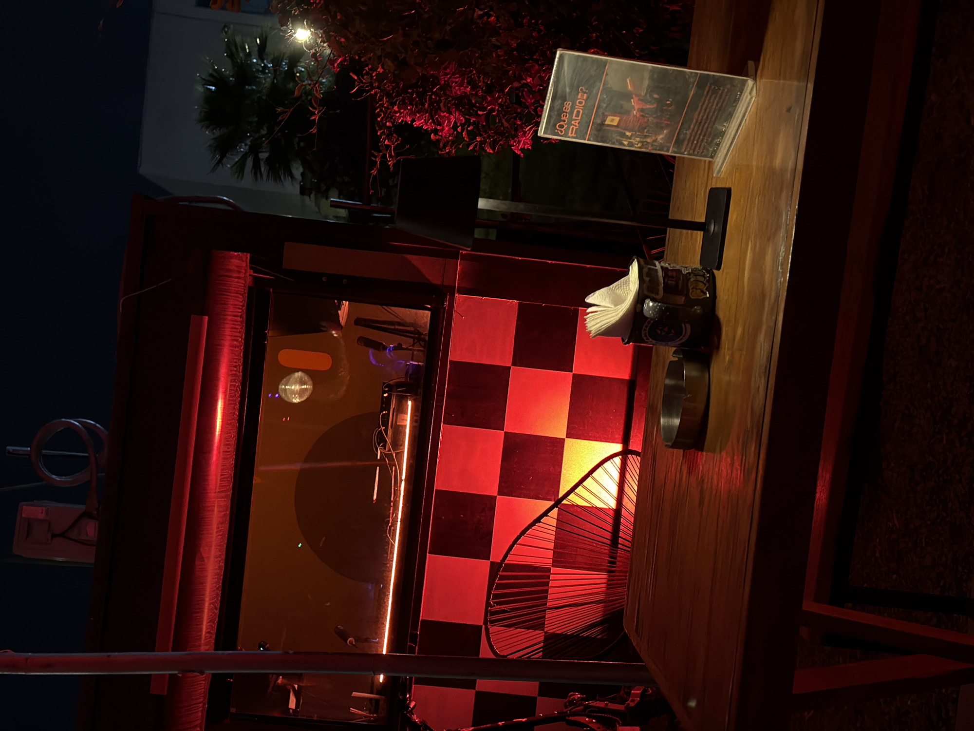 The image depicts an outdoor setting at night, likely a bar or eatery in San Andrés Cholula, Mexico. A wooden structure with a checkered red and black pattern serves as a partition. Inside the structure, there is a circular object illuminated by an orange light, resembling a speaker. On top of the structure, there is a wooden table with a sign titled "¿QUE ES RADIO?" (What is Radio?), a dispenser with napkins, and an ashtray. The overall lighting is dim with reddish tones. In the background, a building and vegetation are visible.