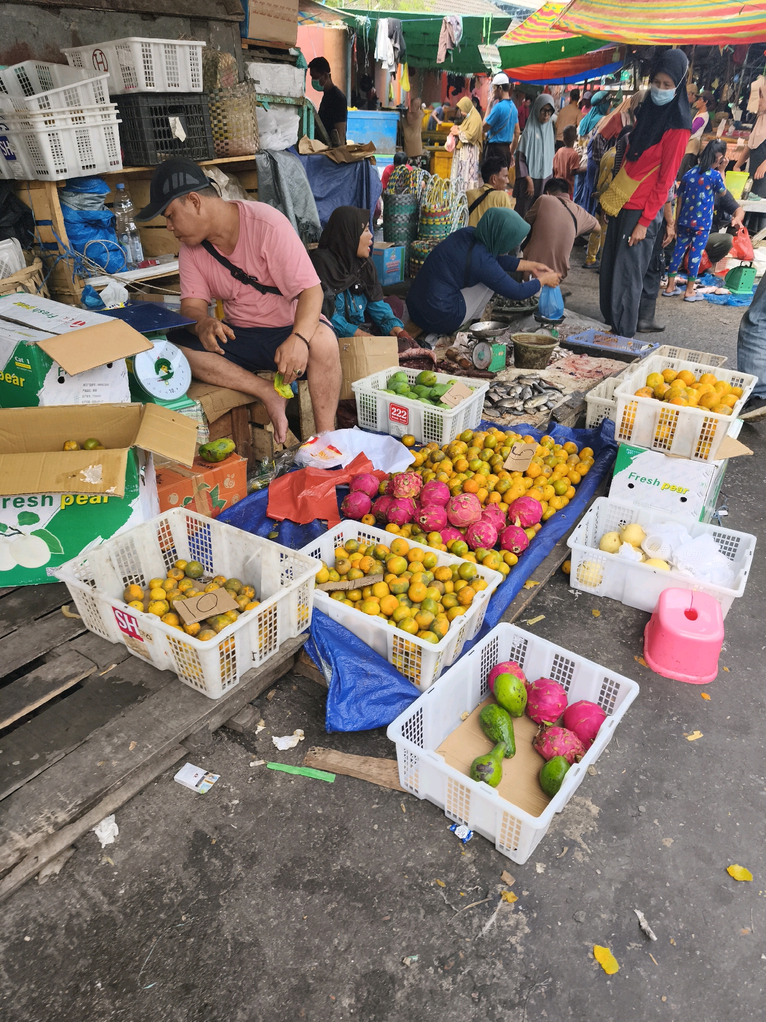 This image depicts a busy outdoor market stall in Palembang, Indonesia, likely specializing in produce. A man wearing a pink t-shirt and a black baseball cap sits on a wooden crate, facing away from the camera. He is examining a yellow plastic bag in his hands. Next to him, a woman in a dark headscarf and blue top is crouched down, appearing to be weighing produce on a green scale.
In the foreground, several white plastic crates are filled with various fruits. One crate prominently displays a handwritten sign with the number "10" on it, filled with small, round oranges. Another crate next to it holds more oranges. Further down, a crate contains dragon fruit and avocados. To the left, green boxes labeled "Fresh Pear" are stacked, with one open to reveal some fruit. Scattered around the crates are discarded leaves, wrappers, and a pink plastic stool.
In the background, the market is bustling with activity. Numerous people, some wearing face masks, are browsing and interacting. Colorful tarps are stretched overhead, providing shade. The ground appears to be a mixture of concrete and dirt, typical of an open-air market. The overall scene conveys a vibrant and active marketplace environment.