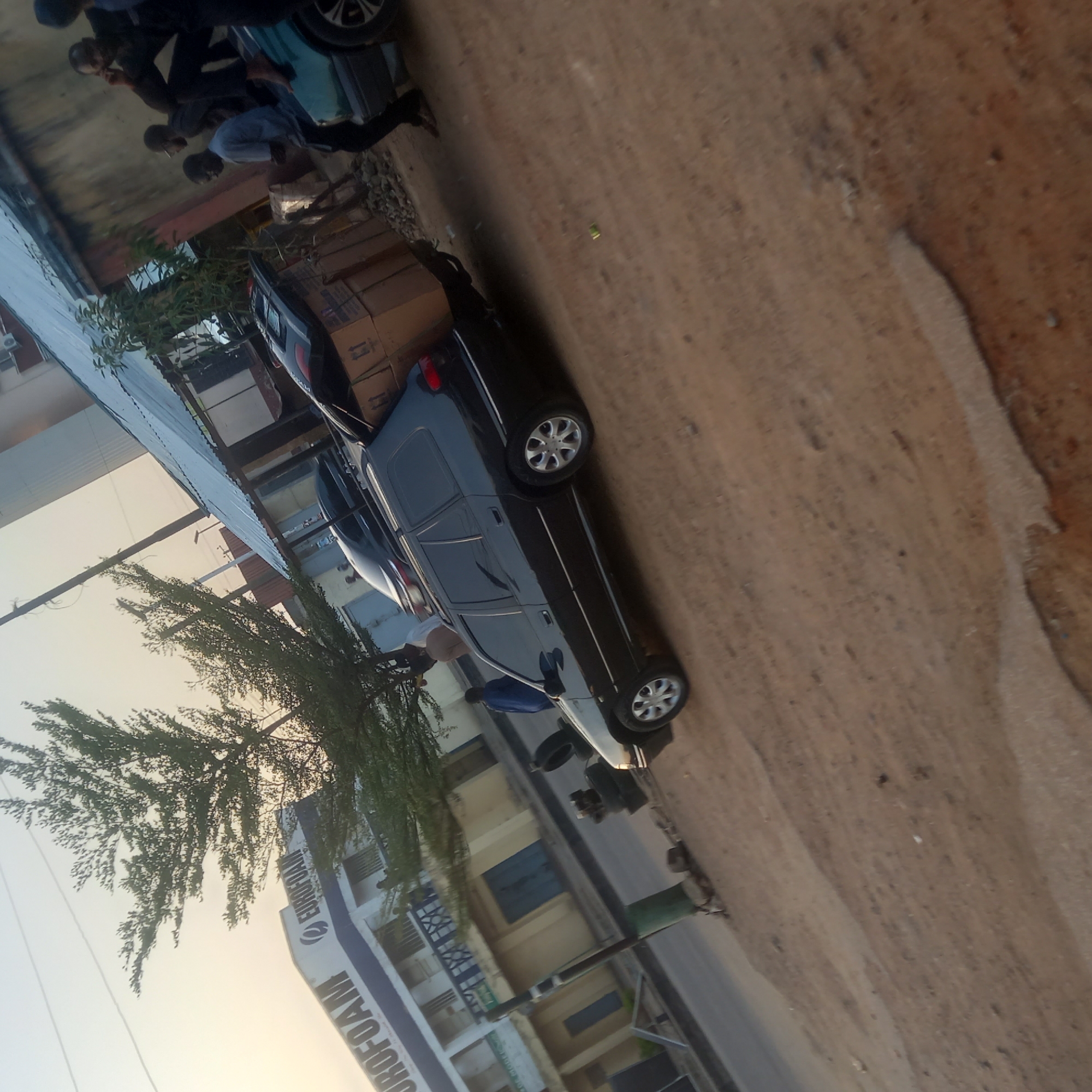 The image shows a black car parked on a dirt road next to a building. Several people are standing near the car, and cardboard boxes are stacked on top of the car. The building is labeled "EUROFOAM". The location is Dadi Riba, Nigeria.