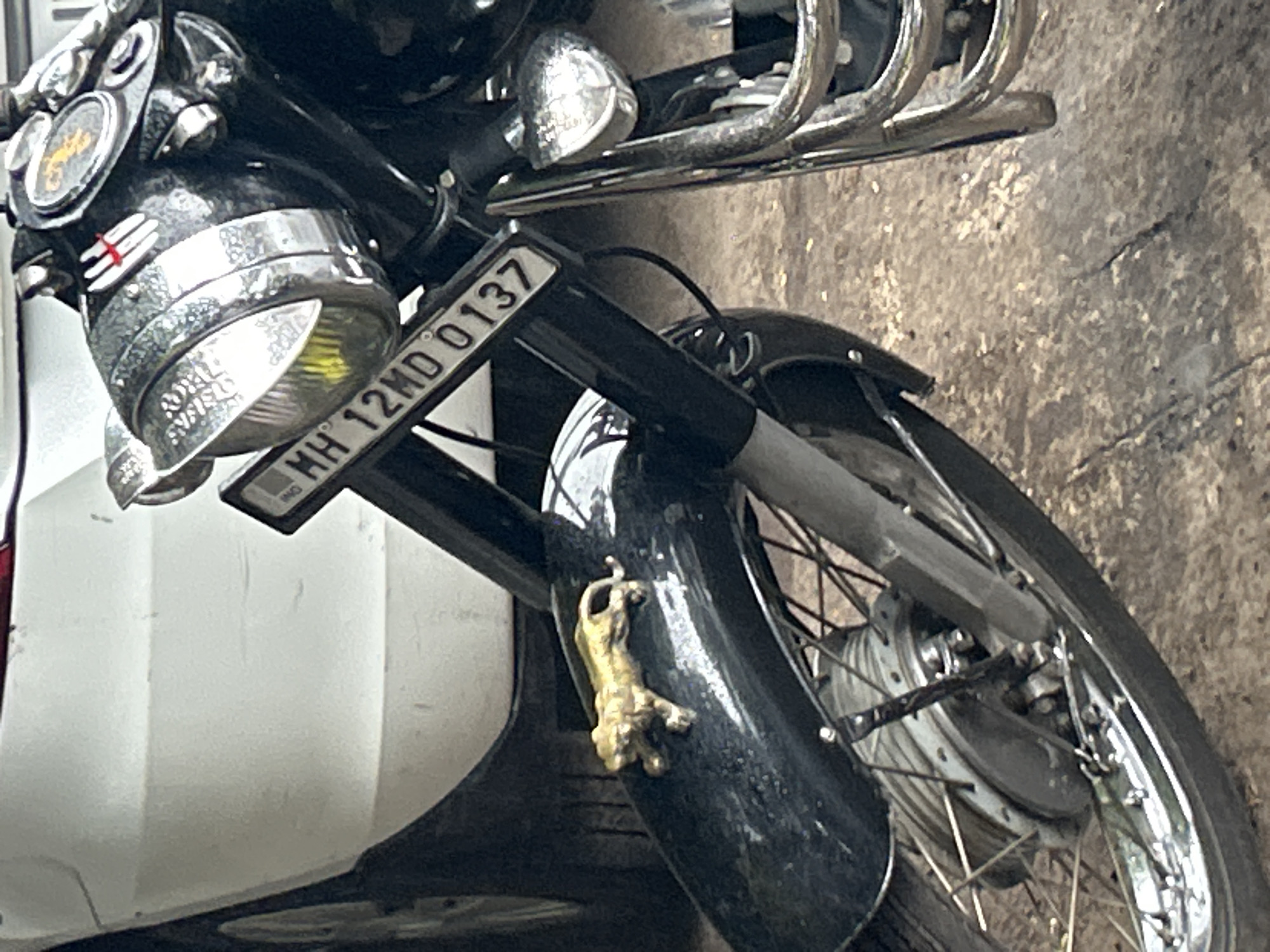 The image shows the front of a black Royal Enfield motorcycle parked outdoors, possibly on a street or driveway, in Pune, India. The motorcycle has a golden figurine of a leopard or tiger attached to its front fender. A license plate, "MH 12MD 0137," is visible on the front of the motorcycle. The motorcycle is partially obscuring the rear of a white car. The ground appears to be asphalt or concrete. The lighting suggests it is daytime.