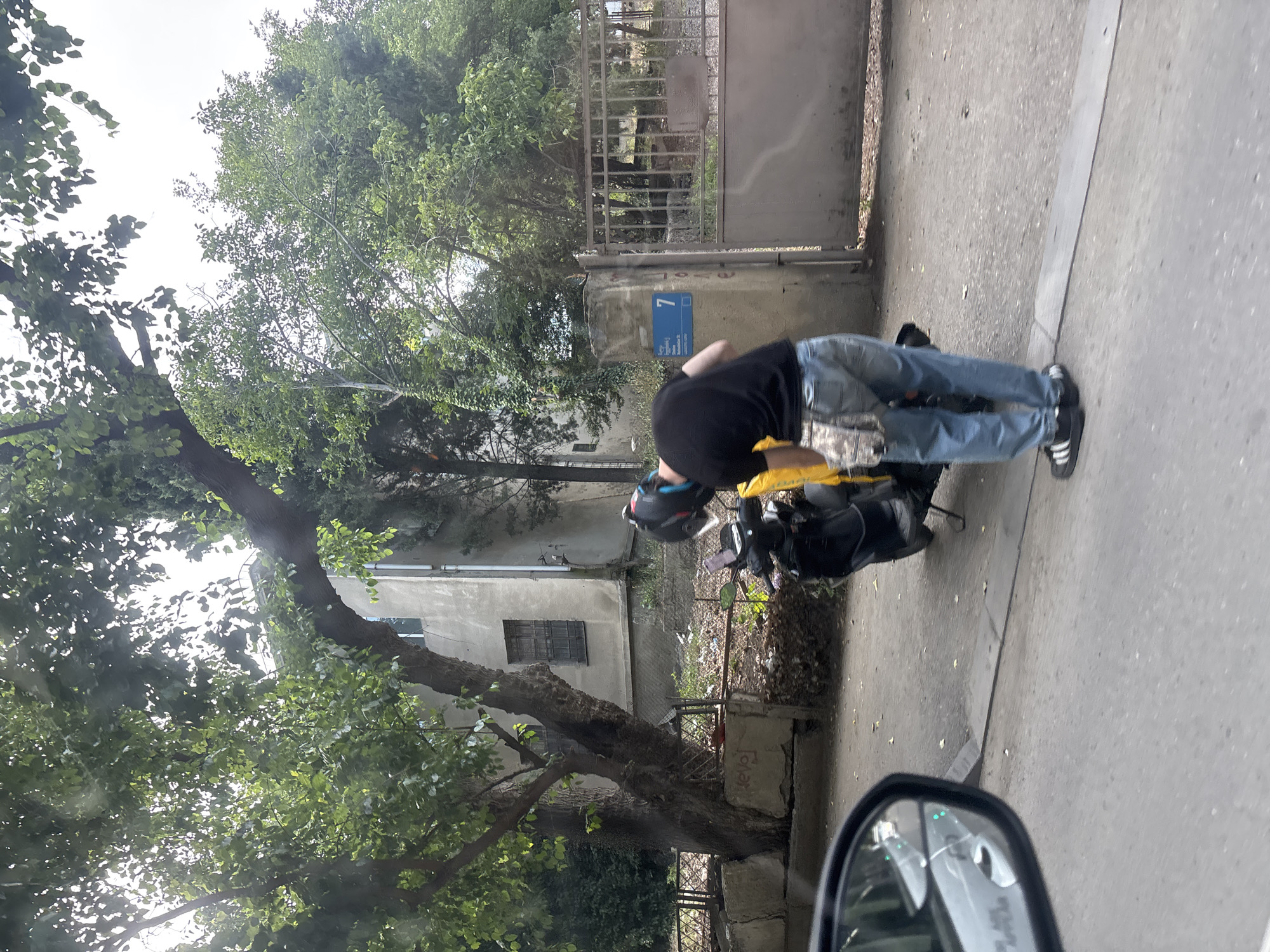The image depicts a person wearing a black t-shirt, blue jeans, and black and white sneakers, bent over a motorcycle on a sidewalk. The person is also wearing a helmet. The motorcycle is black and has a yellow bag attached to the back. The scene is set in an urban environment with a building in the background, trees, and a metal fence. A street sign with the number "7" is visible on a concrete pillar. The ground is paved with asphalt. The image appears to be taken from a vehicle, as a car's side mirror is visible in the bottom right corner. The lighting suggests it is daytime, and the presence of green leaves on the trees indicates it is likely spring or summer. The overall atmosphere is neutral, with no strong emotional cues. The visible text on the street sign is "7" and some other writing in a foreign language that is difficult to decipher.