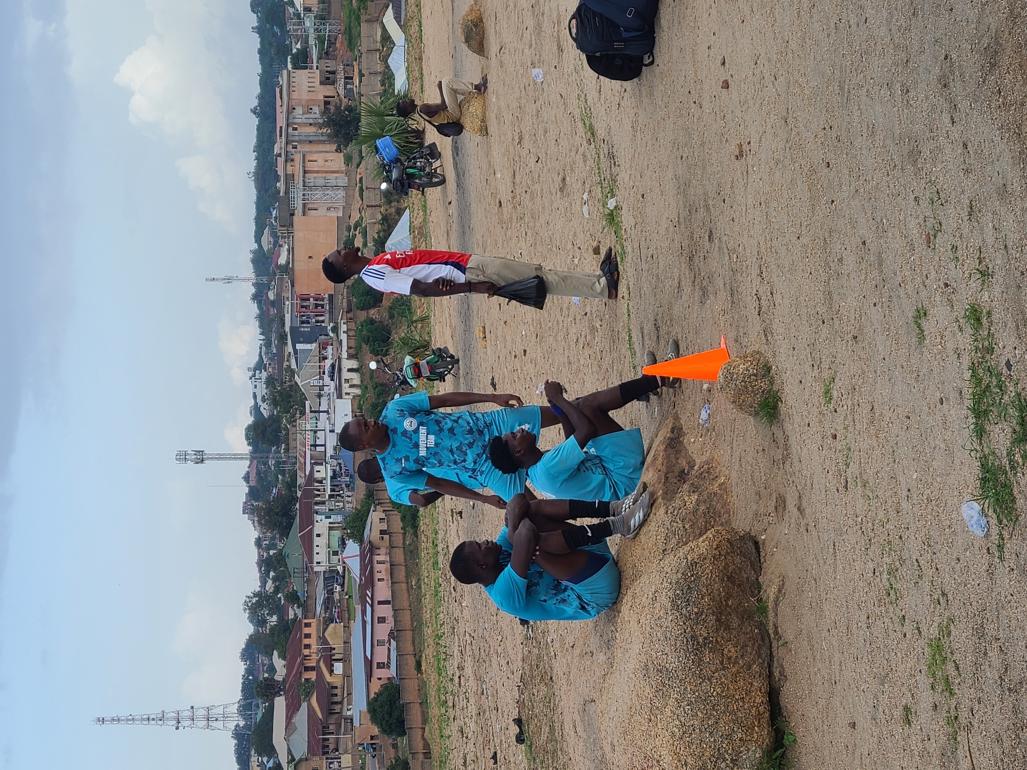 The image depicts a group of young men in what appears to be an outdoor training session in Jos, Nigeria. The scene is set on a sandy, uneven terrain with scattered rocks and sparse vegetation, overlooking a densely populated urban area with buildings and communication towers. The time of day seems to be late afternoon, given the soft lighting and shadows.
There are at least five individuals visible. Three of them are wearing matching light blue athletic wear with a camouflage-like pattern and text that reads "MOVEMENT FEMI". One of these individuals is sitting with his knees pulled up to his chest, another is lying down with his legs bent, and the third is standing. Another person, dressed in a red and white t-shirt and beige trousers, is standing and looking upwards. A fifth person is partially visible at the bottom right corner of the frame. Two motorcycles are parked in the background, and an orange traffic cone is positioned on the ground, suggesting a training drill or practice session is underway.
The backdrop reveals a sprawling cityscape with various architectural styles, from modern buildings to more traditional structures, interspersed with trees. The sky is partly cloudy, indicating a temperate weather condition. The overall mood appears to be one of focus and physical exertion, typical of a sports training environment. The presence of the urban landscape in the background provides a clear geographical context for the location.