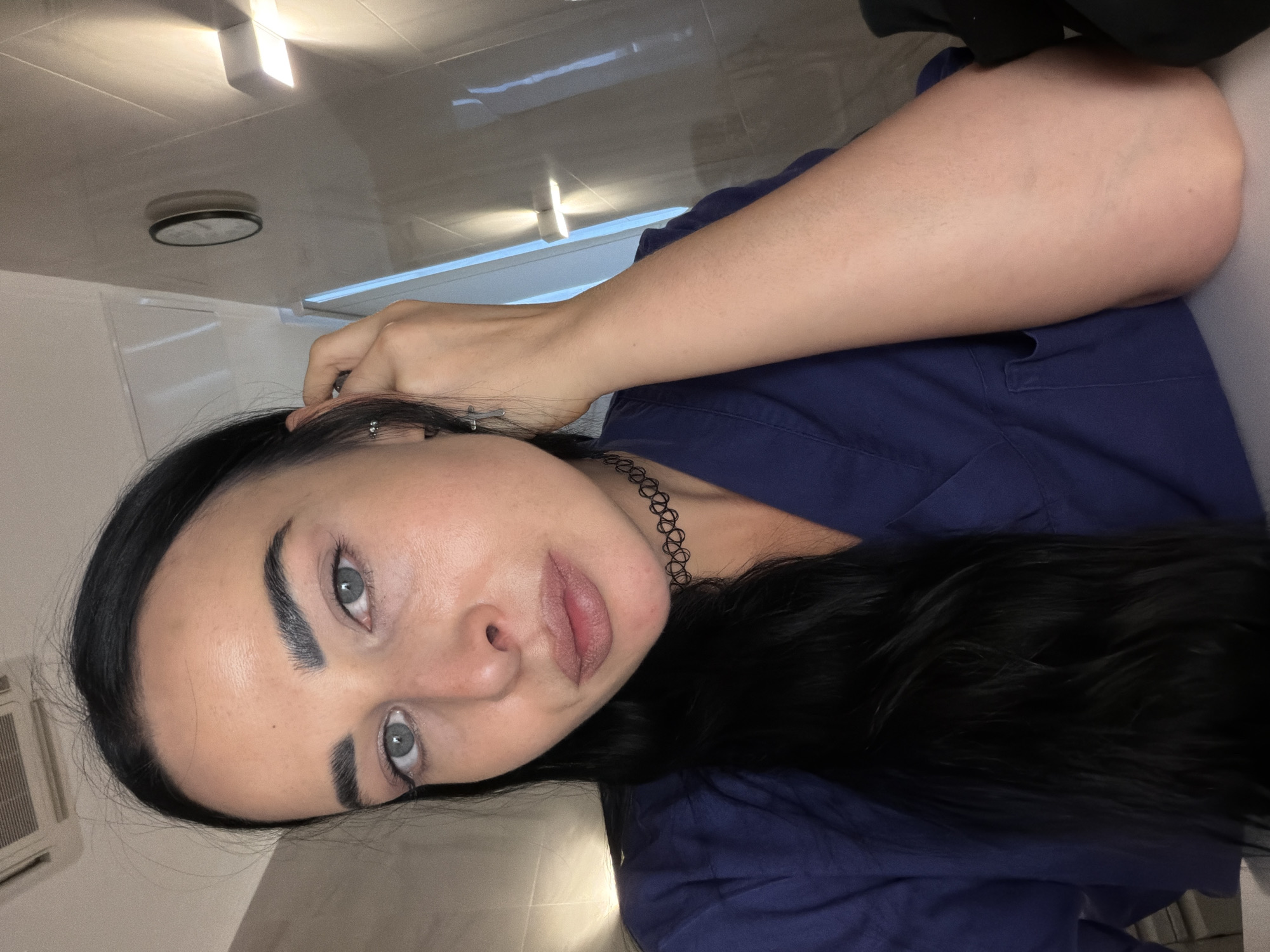 The image is a portrait of a young woman with long, dark, wavy hair. She has fair skin, prominent dark eyebrows, and striking blue-green eyes that gaze directly at the camera. Her lips appear naturally colored. She is wearing a dark blue top, possibly a scrub top or uniform, and a black, patterned choker necklace around her neck. Several small, silver-toned earrings are visible in her left ear. Her left hand is raised, with fingers gently touching her hair near her temple.
The woman is posing for a selfie or a close-up photograph, indicated by her direct gaze and the tilted, unconventional angle of the shot, which suggests she might be holding the camera herself or lying down. Her expression is calm and direct, without a strong smile.
The setting is an indoor space, characterized by bright, artificial overhead lighting. The background features light-colored walls and a ceiling that appear to be tiled or panelled, reflecting the light. Two modern light fixtures are visible on the ceiling: one square and one round. A partial view of what seems to be an air conditioning vent or similar unit is present in the lower left background. The overall impression is a clean, well-lit, functional interior, possibly a bathroom, clinic, or office in Saint Petersburg, Russia.
There is no visible text in the image.