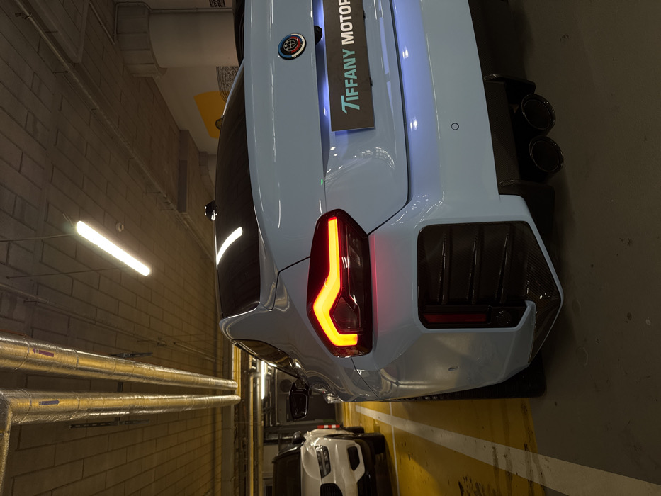 A light blue BMW M2 (G87 generation) is parked in a designated bay within an underground showroom or parking facility belonging to Tiffany Motor in Warsaw, Poland. The vehicle is viewed from a rear three-quarter perspective, with its left LED taillight illuminated in red. The rear bumper incorporates a prominent dark diffuser with vertical fins, likely made of carbon fiber, and features quad dark-finished exhaust tips. A rectangular panel below the trunk lid, illuminated by blue light, displays the text "TIFFANY MOTOR." The BMW emblem is present on the trunk lid. To the immediate left, the front portion of a white SUV, consistent with a Volvo, is partially visible. The environment consists of a concrete floor marked with yellow parking lines, a concrete ceiling with exposed silver-colored HVAC ducting, and multiple overhead fluorescent tube lights providing illumination. White concrete walls and pillars are visible in the background. No individuals are present.