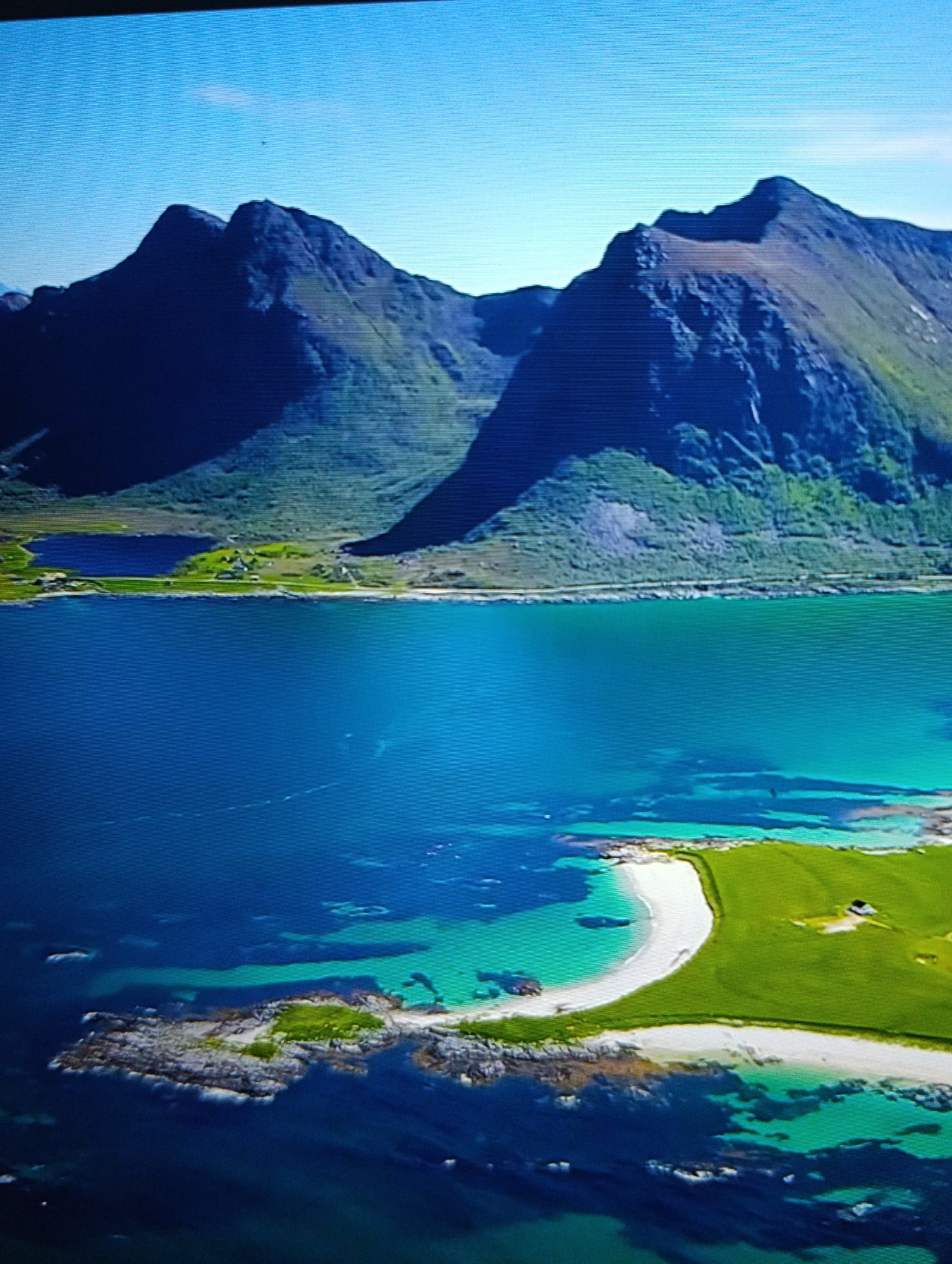 The image depicts a coastal scene with mountains in the background. The mountains are steep and rocky with some vegetation. In front of the mountains is a body of water, likely a fjord or bay. The water transitions from a deep blue color to a lighter, turquoise hue closer to the shoreline. A small island is visible in the foreground, featuring a white sandy beach, green grassy areas, and rocky outcroppings. There appears to be a small structure on the grassy area. The sky is clear and blue. The location is unknown.