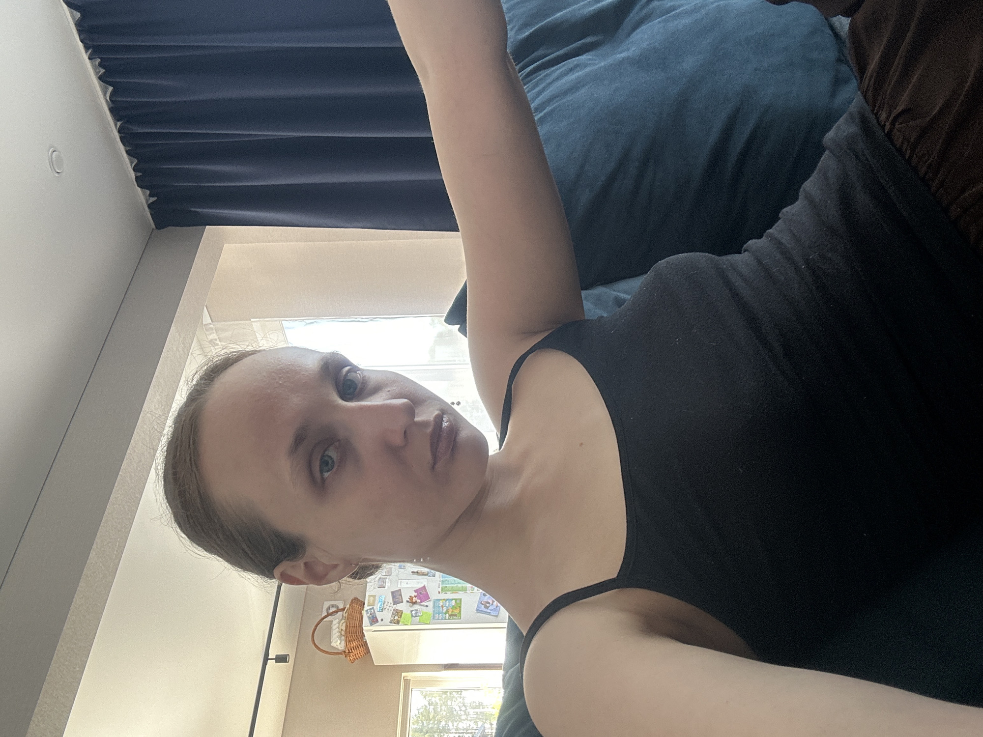 A woman is lying on a blue cushion, wearing a black tank top and brown clothing. She is indoors, and a curtain is visible in the background. A window shows the outdoors. A refrigerator is visible in the background. There is no direct evidence indicating this image was taken in Sepych, Russia.