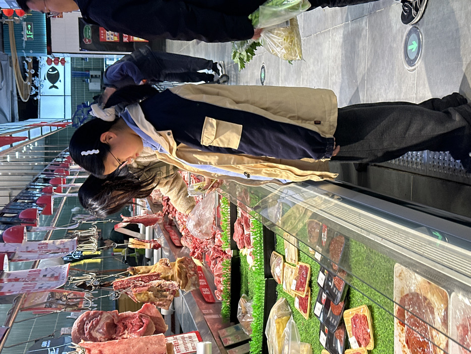 The scene depicts the interior of a supermarket's meat and deli section in Jiangjin, China. In the foreground, a young female, appearing to be a teenager, stands in profile, looking intently into a refrigerated glass display case. She has dark hair styled in a ponytail with a white hair clip, wears glasses, a light blue hooded top under a beige and navy-blue two-tone jacket with a utility pocket on the left arm, and dark grey sweatpants. Directly behind her, an adult female with long dark hair, wearing a light brown sweater, actively selects raw meat from a bulk display using a plastic-gloved hand, while holding a small, clear package.
The meat display spans the length of the counter, featuring various raw meat cuts; some are hanging from hooks above, while others are piled loose or packaged in clear plastic and black trays, arranged on artificial green grass linings. Price tags, including ones marked "10.8" and "15.8", hang above the bulk meat. To the far right, a person is partially visible, holding a clear plastic bag containing bean sprouts and other green leafy vegetables. The store's environment includes bright overhead lighting, accentuating red lights, and a concrete floor with a green arrow directional marker. In the background, a sign with a fish emblem and "1F ->" is visible, alongside decorative red lanterns. Additional customers are indistinctly present in the deeper background.