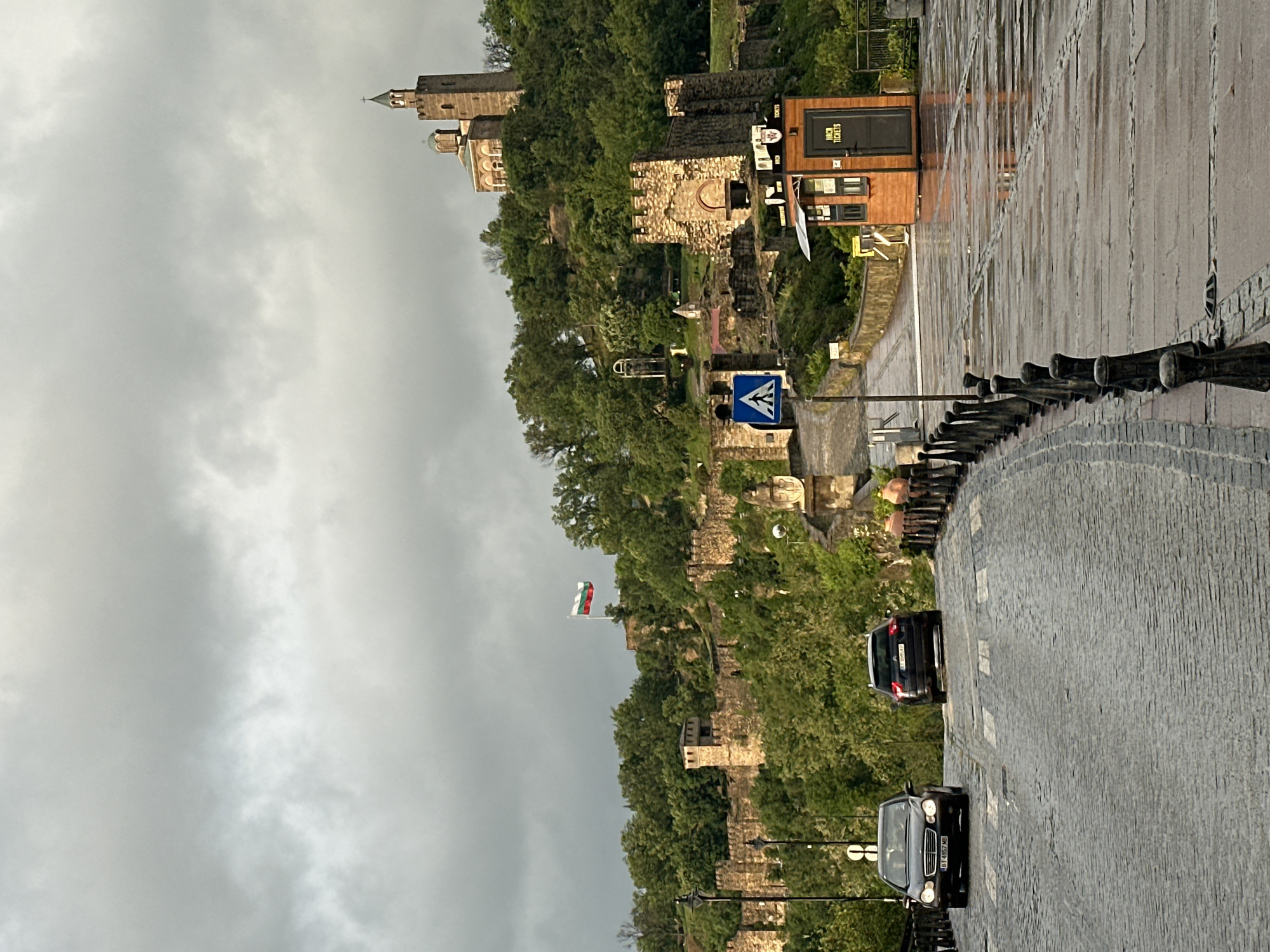 The image depicts a street in Veliko Tarnovo, Bulgaria, likely near the Tsarevets Fortress. The scene is overcast, possibly rainy, as the cobblestone street is wet and reflective. A section of the fortress wall covered with lush greenery runs along the left side of the street. The Bulgarian flag is seen waving near the fortress wall. Two cars are traveling on the street. Pedestrian crossing sign is visible alongside the road. In the distance, the fortress buildings can be identified on top of the hill. A small ticket booth sits on the right side of the road. The street is lined with a short metal barrier.
