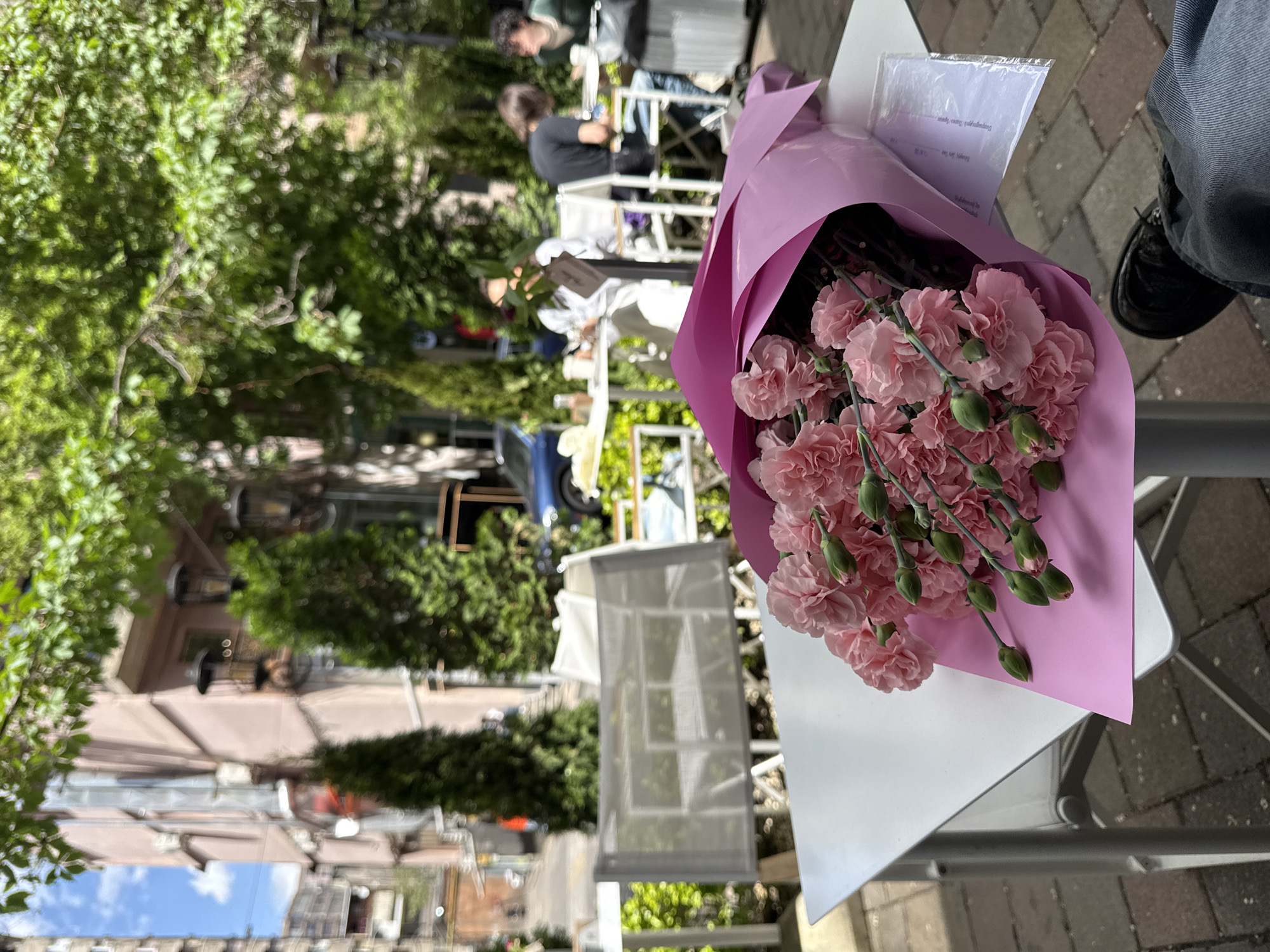 The image depicts a bouquet of pink carnations wrapped in pink paper on a white table, likely an outdoor cafe setting in Yerevan, Armenia. Visible in the background are trees, buildings, and people seated at other tables, suggesting a street-side location. A person's leg wearing jeans and a black shoe is also visible in the right part of the frame. A white paper is tucked into the flower bouquet. The scene is captured in daylight.