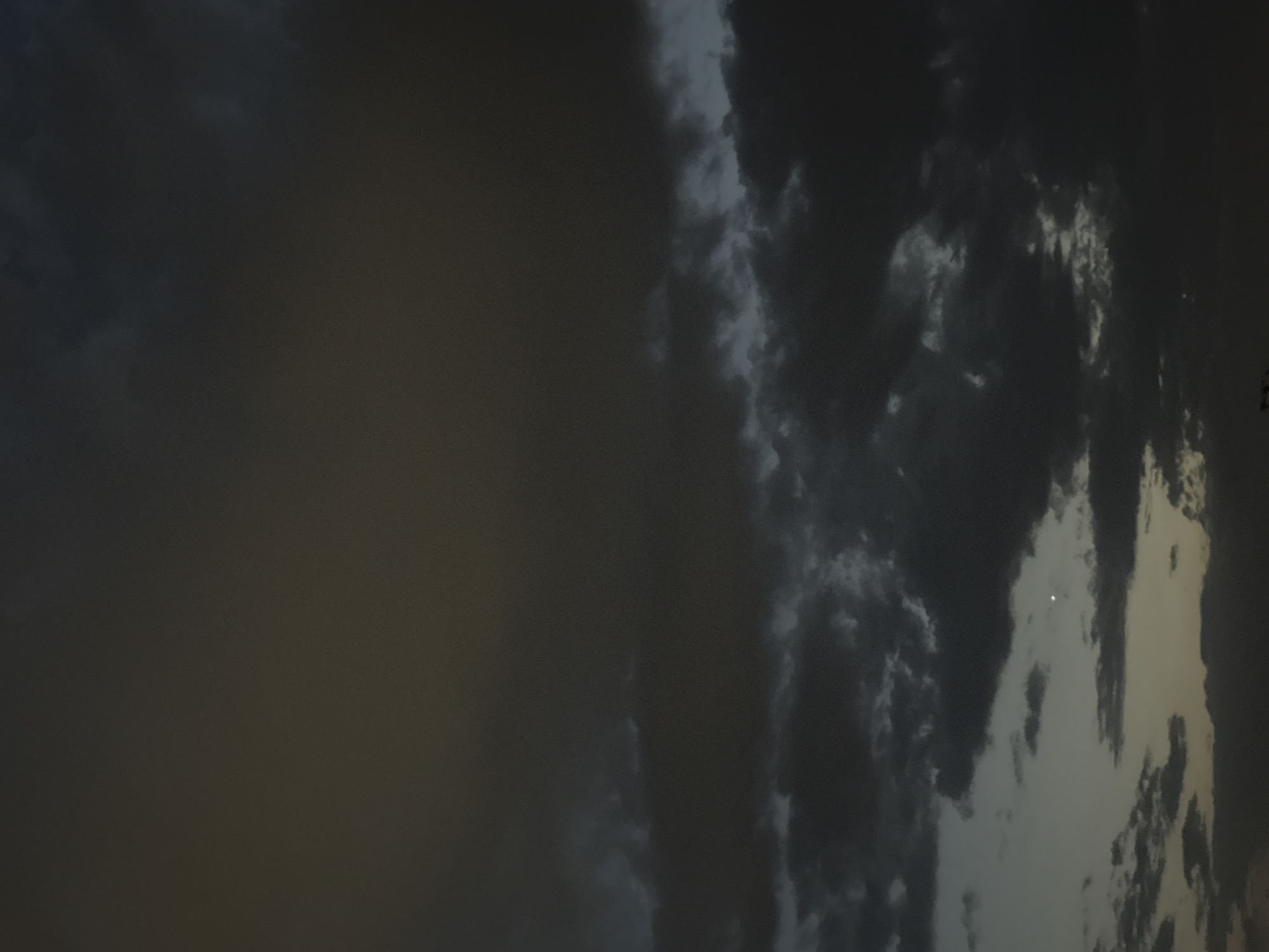 A nighttime image captures a sky filled with dark, heavy clouds. The clouds are dense and layered, with some thinner patches allowing a faint glow from the horizon to appear as a pale yellow-gray hue. A small, bright point of light, possibly a distant planet or star, is visible in the lower middle section of the image, surrounded by dark cloud formations. The overall atmosphere is somber and overcast, with the lower edge of the frame showing the silhouette of unseen objects, suggesting the presence of the city of Bishkek, Kyrgyzstan, at ground level.