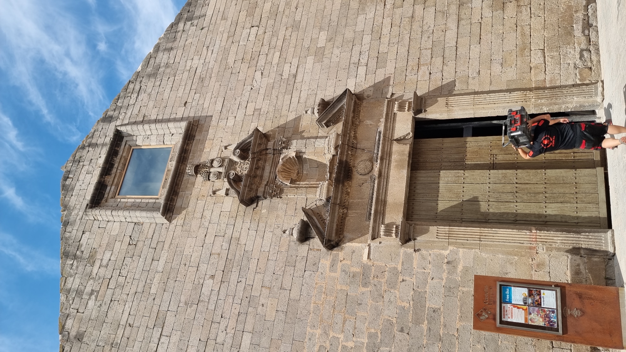 The image shows the entrance to a stone building, likely a historic church or castle, in Castelló d'Empúries, Spain. The building's facade is constructed from light-colored stone blocks. Above a large set of wooden double doors, there is an ornate stone architectural feature with decorative carvings, a small niche, and a crown-like element. A single window with a wooden frame is visible higher up on the facade. To the left of the entrance, a brown informational stand with a poster displaying event information and bus routes is present. In the foreground, a person is walking away from the viewer towards the open doorway, carrying a black and red toolbox or case on their head. The person is wearing a black t-shirt with a circular red logo and black shorts with red accents. The ground in front of the building is paved with light-colored stones. The sky above is blue with scattered white clouds.