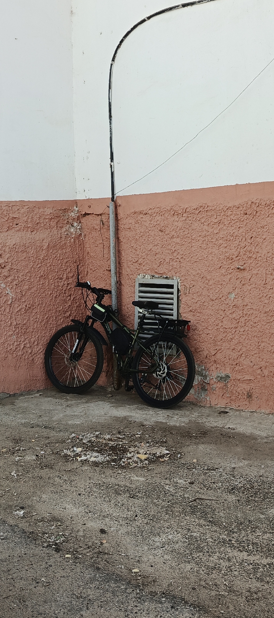 A dark green mountain bike is parked diagonally in a corner formed by a textured salmon-colored wall and a white wall. A black cable runs from the top of the frame down along the white wall, curving over the salmon wall. The bike has black wheels with silver spokes, a black frame, black handlebars with a small green and yellow pouch attached, and a black seat. Behind the bike, mounted on the salmon wall, is a white louvered vent. The ground is a rough, gray concrete surface covered in debris, including small pieces of paper and dirt. The scene appears to be an outdoor, urban environment.