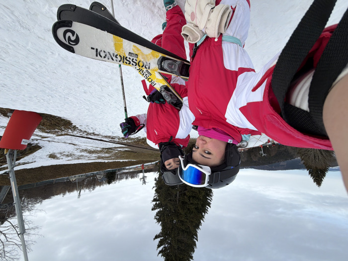 Two individuals stand on a snow-covered ski slope in what appears to be a resort setting, potentially in Belmont, France. The person on the left, foreground, is taking a selfie, with her right arm extended and the camera pointed towards herself and the second individual. She wears a red and white ski suit, a black helmet with blue mirrored goggles pushed up, a pink neck gaiter, and light-colored gloves tucked into her light blue belt. The second individual, positioned slightly behind and to the right, also wears a matching red and white ski suit, a black helmet, black goggles, and a black balaclava covering her lower face. She holds two ski poles in her right hand and carries a pair of "ROSSIGNOL" brand skis, yellow, white, and black, over her left shoulder. The ski slope exhibits patches of snow interspersed with visible brown earth/grass. In the background, pine trees, bare deciduous trees, and distant hills or mountains are visible under an overcast sky. Elements of a ski lift, including metal poles and red safety padding, are present on the right side of the frame.