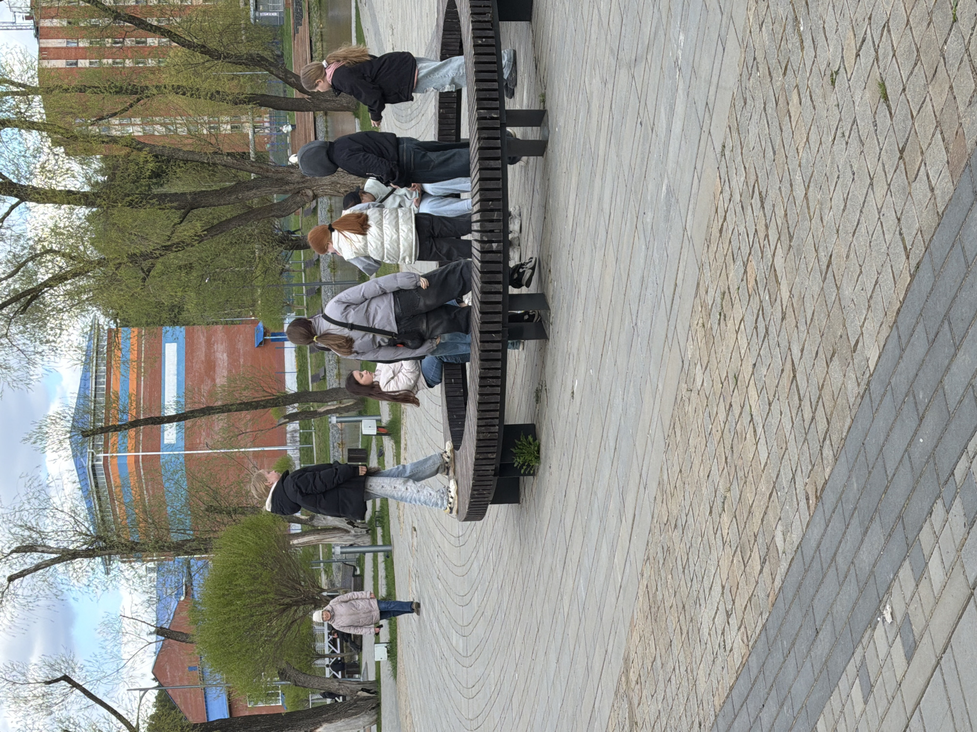 The image depicts a group of six people sitting on a uniquely designed park bench in Votkinsk, Russia. The bench is constructed with vertical wooden slats connected to circular metal supports, resembling a gear. The ground is paved with interlocking brick patterns, and trees with sparse green foliage are visible in the background. A multi-story building with red, orange, and blue horizontal stripes is also present in the distance. One individual is walking away from the bench along the pathway. The weather appears to be overcast.