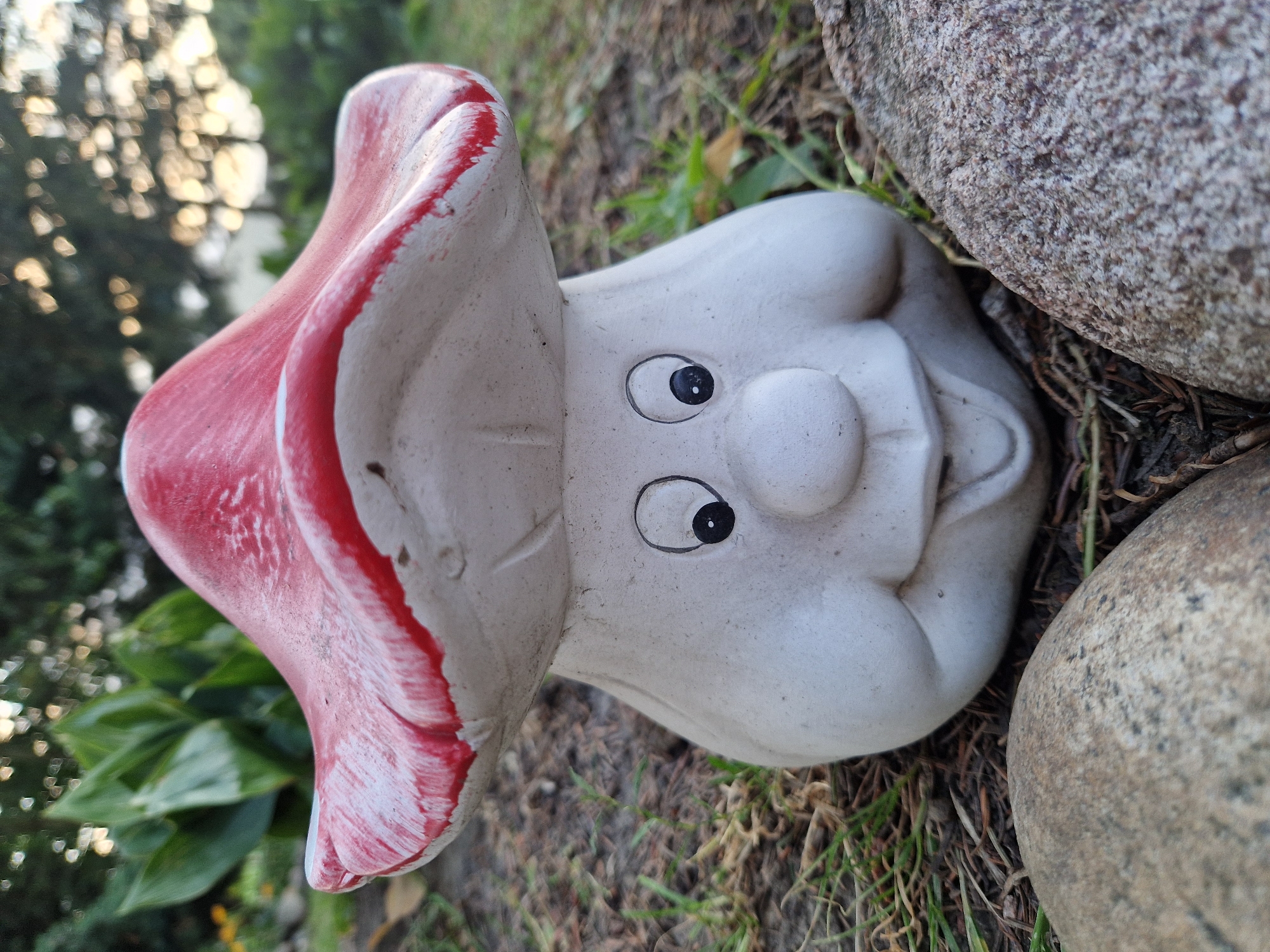 A garden gnome shaped like a cartoon mushroom with a red cap and a white stem with a face is positioned outdoors on the ground. The gnome has black dot eyes, a round nose, and a smiling mouth. The stem of the mushroom has prominent cheeks. The gnome is surrounded by green grass, dry leaves, and mulch. Two large rocks are in the foreground, one on the left and one on the right, partially obscuring the gnome. In the background, there are green leafy plants and trees with blurred foliage and a bright sky.