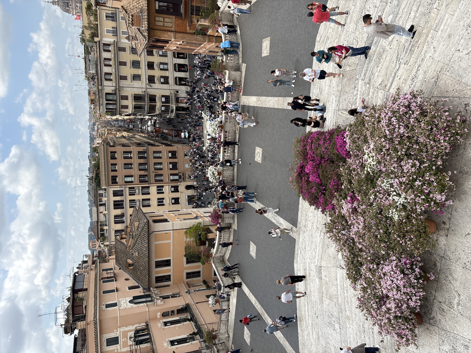 The image presents a vibrant, high-angle view looking down the Spanish Steps in Rome, Italy. In the immediate foreground, a broad section of the iconic stone steps is visible, extensively decorated with large potted flowering plants, predominantly in shades of pink, purple, and white blossoms. Numerous people are scattered across these steps, casually dressed, with some sitting, others standing, or moving about, enjoying the popular landmark.
Beyond the steps, a long, bustling street extends into the distance, flanked on both sides by rows of classic Roman buildings. These multi-story structures feature warm color palettes of cream, ochre, and light pink, with numerous windows and balconies. The street below is also populated with people and a few vehicles are discernible further down. In the far background, the distinct dome of a prominent church, likely St. Peter's Basilica, rises above the city's rooftops.
The scene depicts a lively daytime setting, likely late morning or early afternoon, under a partly cloudy blue sky with bright sunlight casting clear shadows. The atmosphere appears relaxed and engaging, typical of a bustling tourist destination. No discernible text is visible within the image.