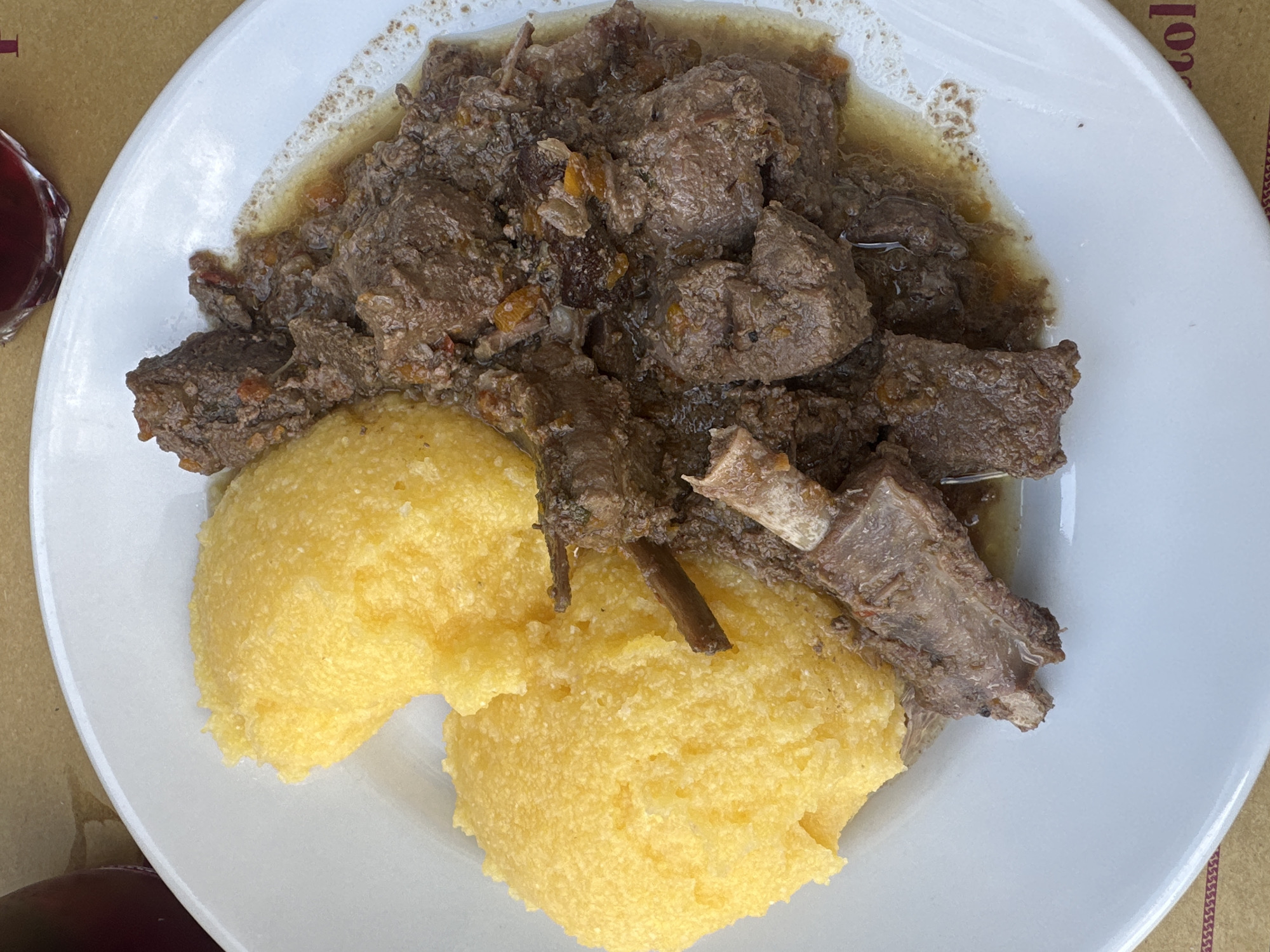 The image displays a close-up, top-down view of a white plate on a light brown, textured surface, possibly a table. The plate contains a portion of a meal consisting of two mounds of bright yellow polenta positioned on the left side of the plate. To the right of the polenta is a generous serving of braised meat in a rich, dark gravy. The meat appears to be cut into bite-sized pieces and includes some bone fragments, suggesting a stew or braised dish. Small flecks of orange, likely from carrots or other vegetables, are visible within the meat and gravy. A small portion of a dark red liquid, potentially wine, is visible in a faceted glass at the top right edge of the frame. The background surface has some faint text or patterns visible, particularly along the bottom edge of the plate. The scene is well-lit, highlighting the textures and colors of the food. The setting appears to be a restaurant or dining establishment in Corniglio, Italy.
