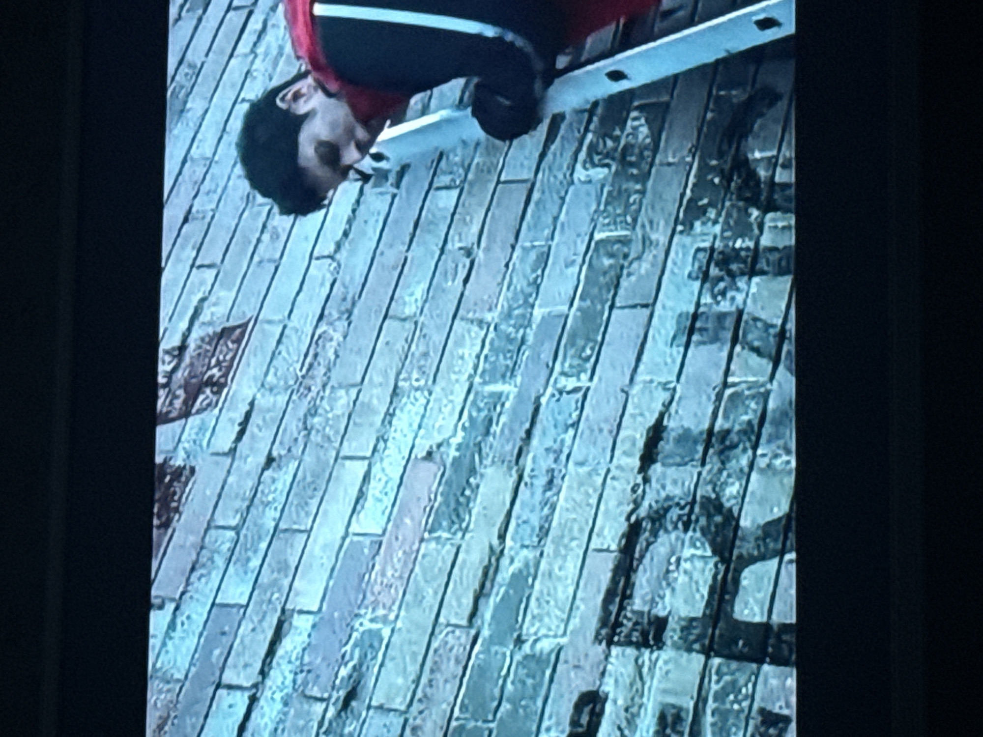 A man is on a ladder leaning against a brick wall. The wall has some graffiti on it. The scene appears to be an outdoor setting. The location is unknown.