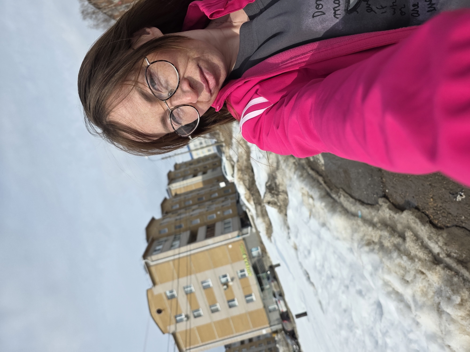 A woman is taking a selfie in front of an apartment building in Votkinsk, Russia. The woman has brown hair, wears glasses, and is dressed in a grey top and bright pink jacket. The apartment building is beige and has multiple floors. There is snow and ice on the ground, indicating a cold environment. The sky is overcast.