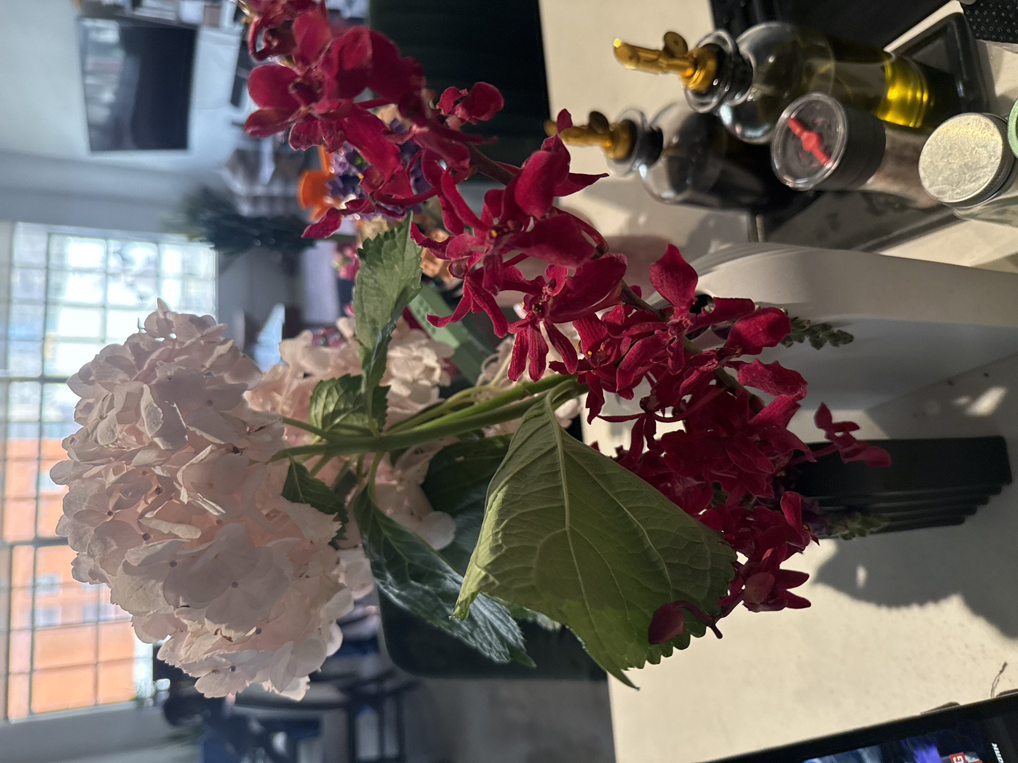 The image shows a bouquet of flowers, predominantly composed of light pink hydrangeas and smaller, vibrant red orchids. A large green leaf is prominently positioned within the arrangement. The flowers are likely in a vase or container, though the base is not fully visible. Behind the flowers, there are glass bottles with metallic dispensers. The background includes a window with multiple panes, revealing a glimpse of a brick exterior and some furniture inside the room. The scene appears to be indoors, and based on the style of the building and the presence of sunlight, it is likely located in a residential or commercial space in Los Angeles, United States of America. A television can also be seen along the bottom right.
