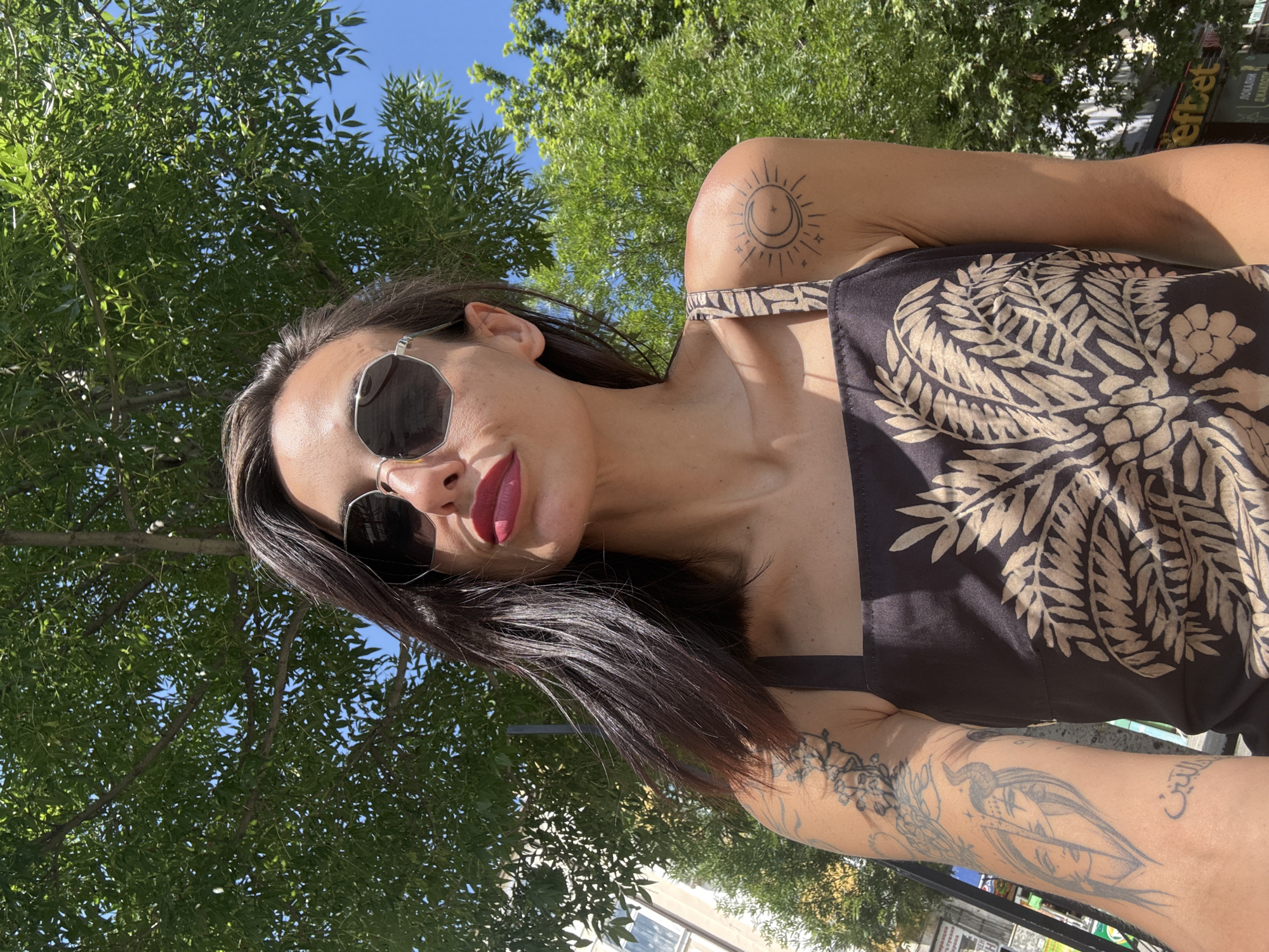 A person, likely female, is captured in a bright, outdoor selfie. She has dark hair, wears large, dark octagonal sunglasses, and vibrant red lipstick. Her attire is a dark top or dress featuring a prominent light-colored botanical leaf pattern.
The subject's right shoulder (viewer's left) sports a tattoo of a stylized sun and crescent moon design. Her left arm (viewer's right) is extensively tattooed with various images, including a horned figure, intricate floral or leafy patterns, and visible Arabic script.
The background is dominated by lush green tree canopies, with patches of clear blue sky visible through the leaves, indicating a sunny daytime setting. The camera angle is low, looking upwards at the subject. In the distance, through the foliage, urban elements are subtly visible. A sign in the background displays Cyrillic text, including "Еfbet" and "ПОКАЗНИ СЛУЖБИ," with "ОНЛАЙН" also faintly discernible, aligning with the Pleven, Bulgaria location context. The overall impression is one of casual confidence, with the person posing directly for the camera.