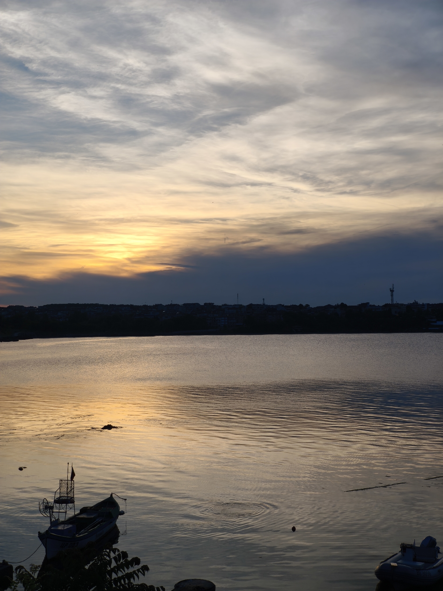 The image depicts a serene sunset over a body of water in Tsarevo, Bulgaria. The sky is a canvas of pastel colors, with hues of orange, yellow, and blue blending together, streaked with wispy white clouds. The sun is setting near the horizon, casting a warm golden glow on the water's surface, which is rippled by gentle waves.
In the foreground, two boats are visible. One is a small, dark-colored fishing boat with a red flag and a metal frame structure, tethered to the shore. The other is a partially visible inflatable dinghy with an outboard motor, floating further out on the water. The shoreline is lined with a silhouetted landscape of buildings, suggesting a town or city.
The overall atmosphere is peaceful and tranquil, capturing the beauty of a coastal sunset in Tsarevo. There are no people visible in the image.