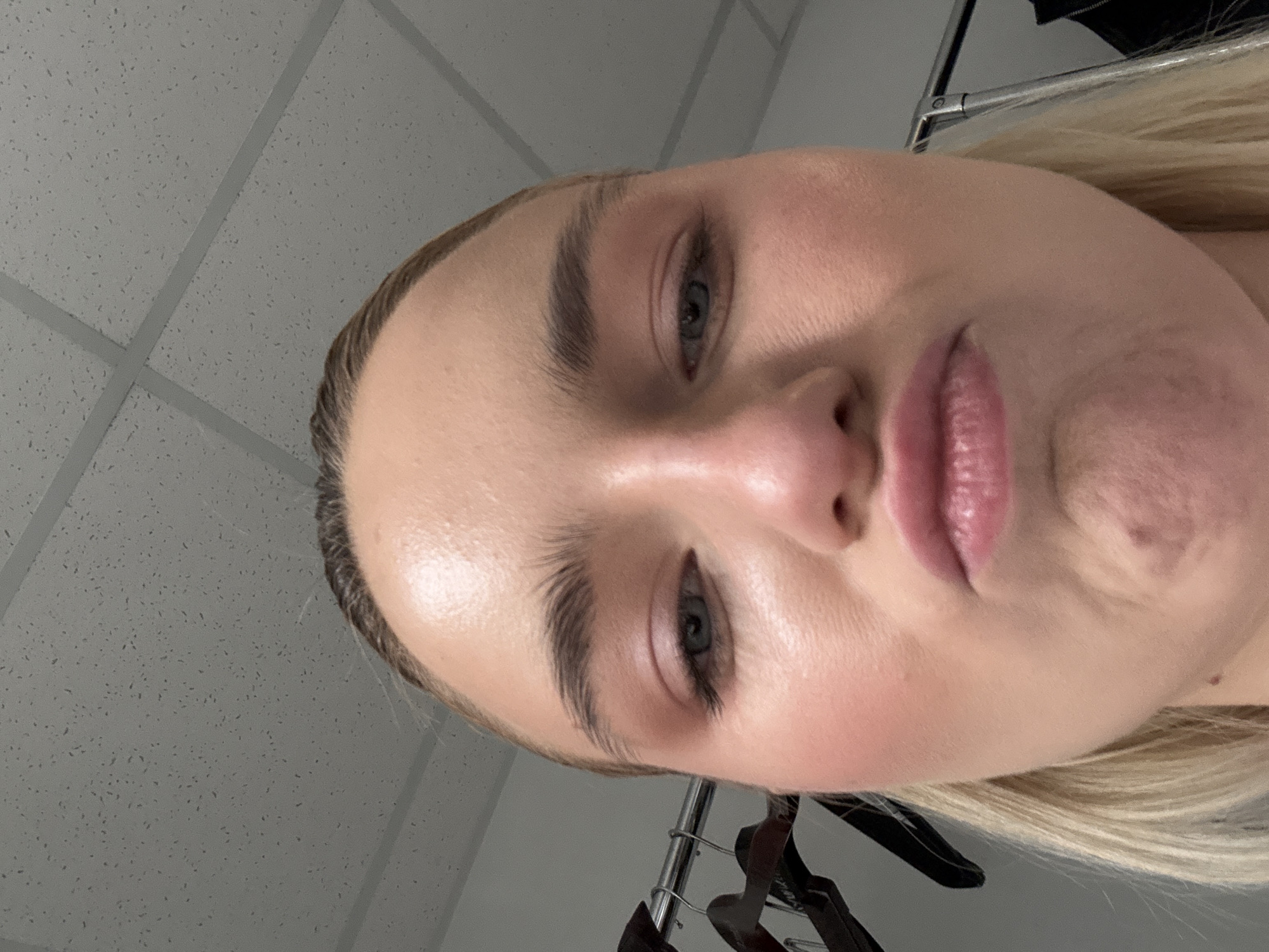 Close-up of a person's face with light skin, light blonde hair, and blue eyes. The person has neutral makeup. The background is a gray textured ceiling and a metal clothing rack with hangers. The setting does not contain sufficient detail to identify the location in Saint Petersburg, Russia.